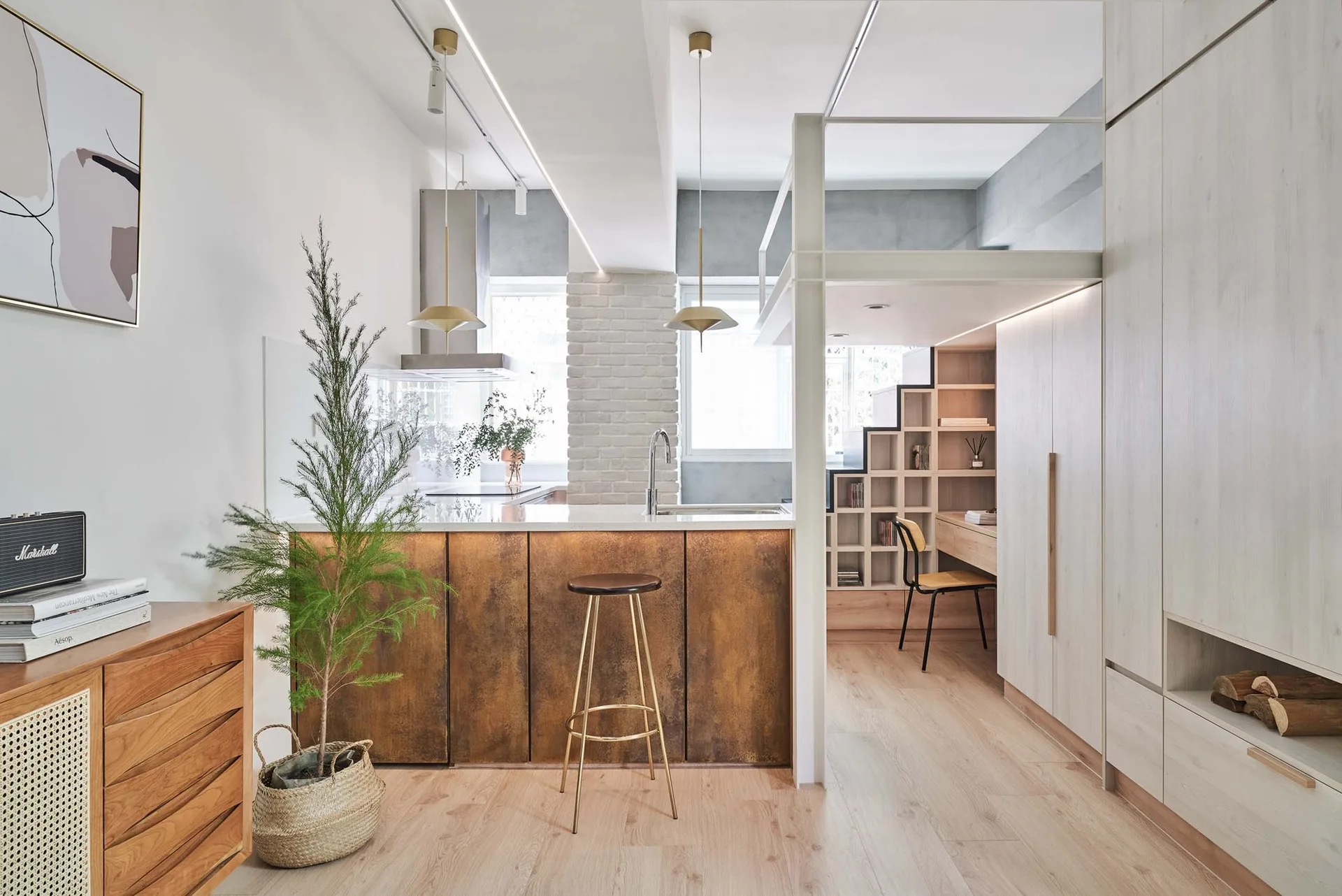 A modern apartment kitchen featuring a rusted metal island under brass pendant lights, alongside a loft workspace with built-in wooden stairs and shelving.