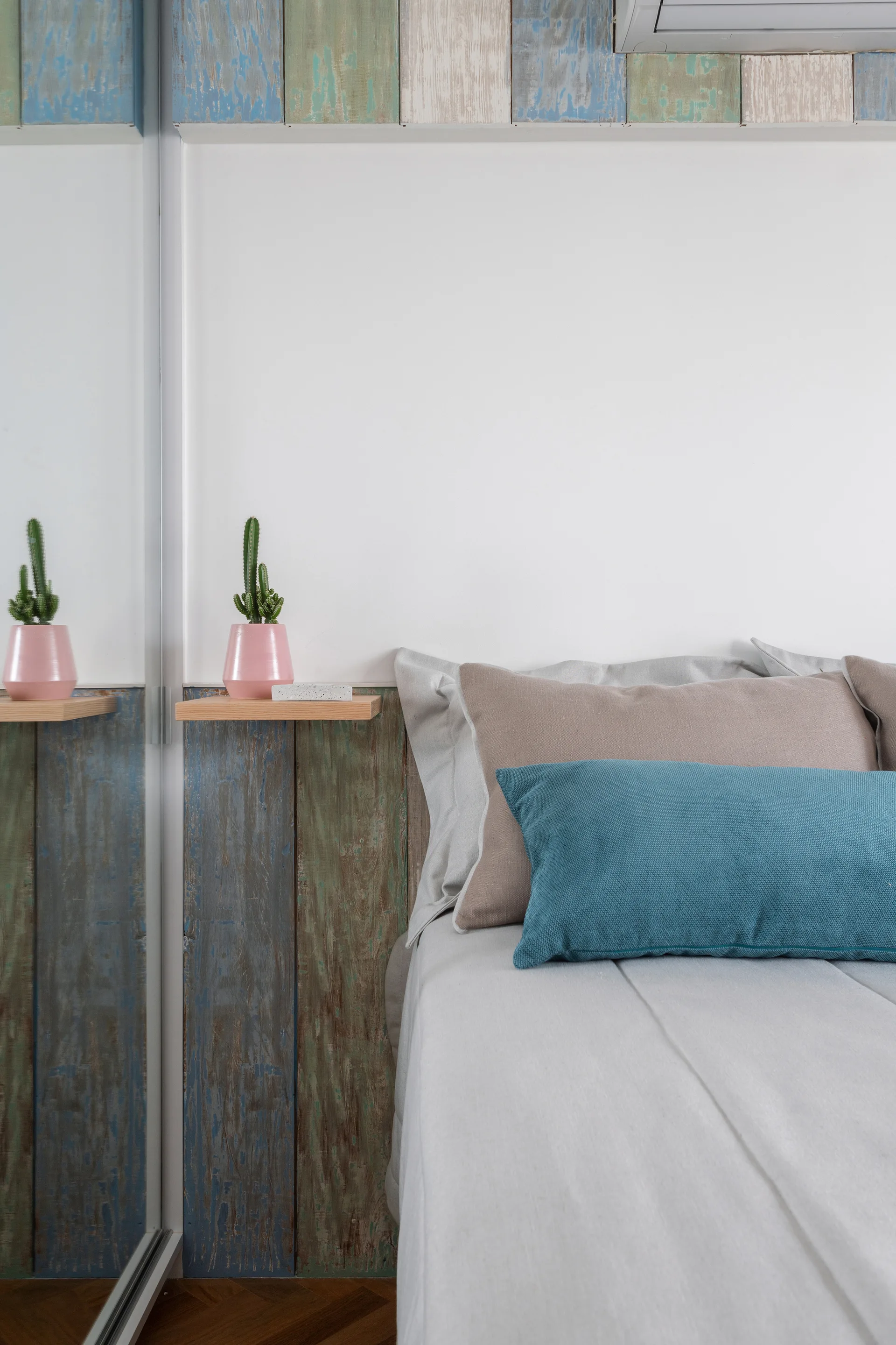 A bedroom bed with teal and beige pillows sits next to a mirrored closet door and colorful weathered wood wall paneling.