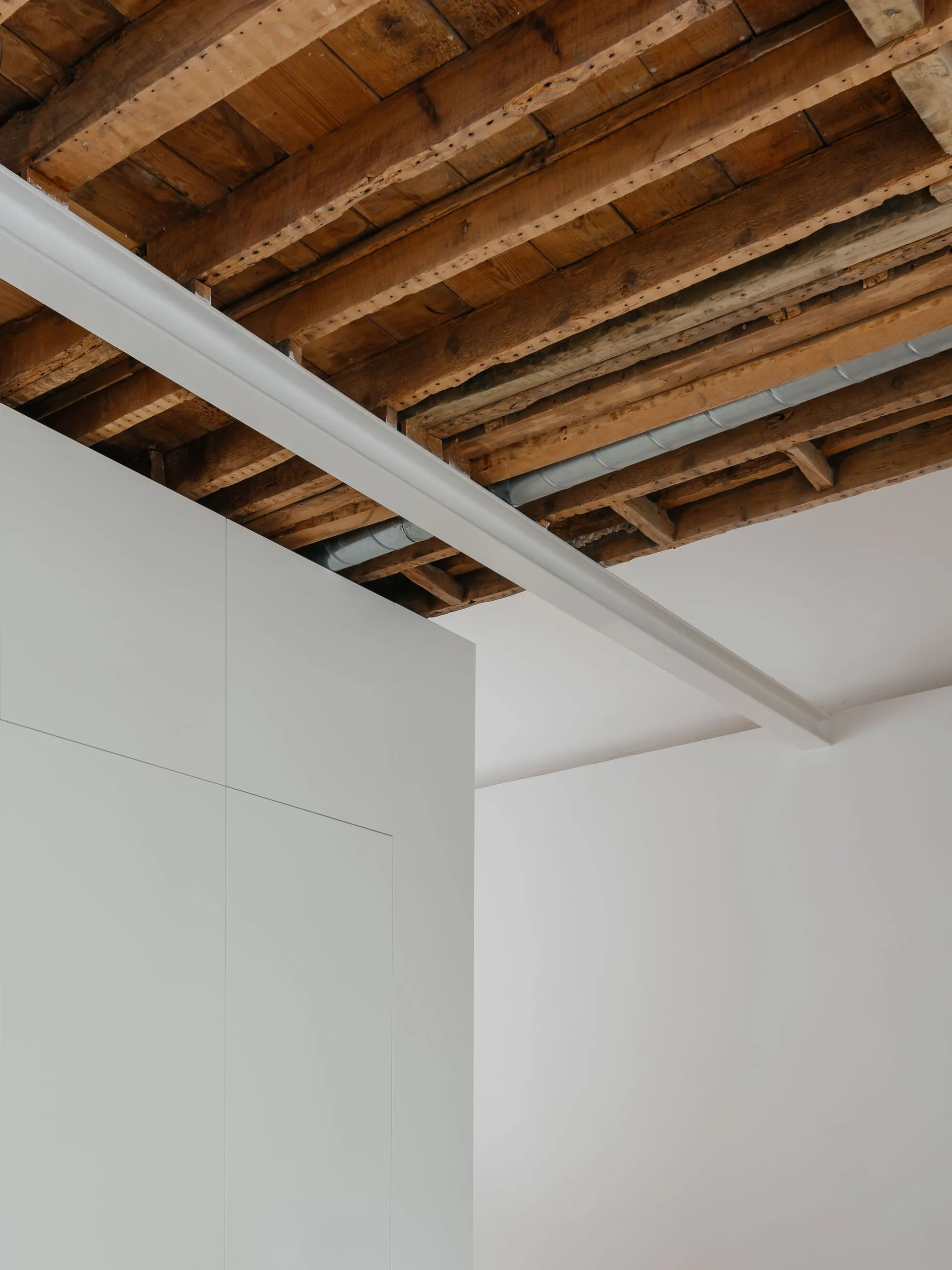A white minimalist partition wall meets an exposed rustic wood ceiling with a white structural metal beam.