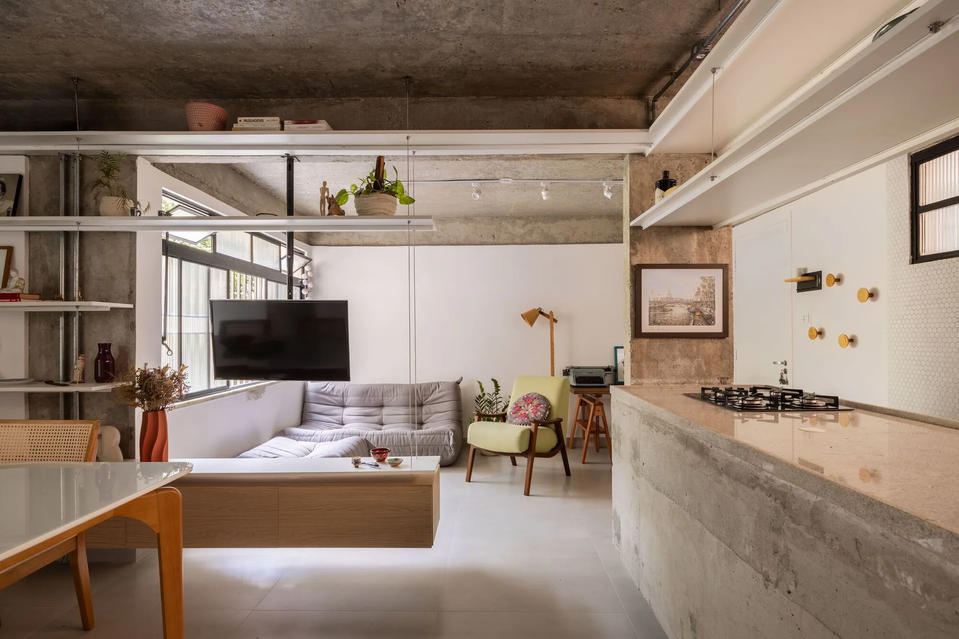 An open plan living area featuring an exposed concrete ceiling, a concrete kitchen counter, a low gray sofa, and suspended white metal shelving.