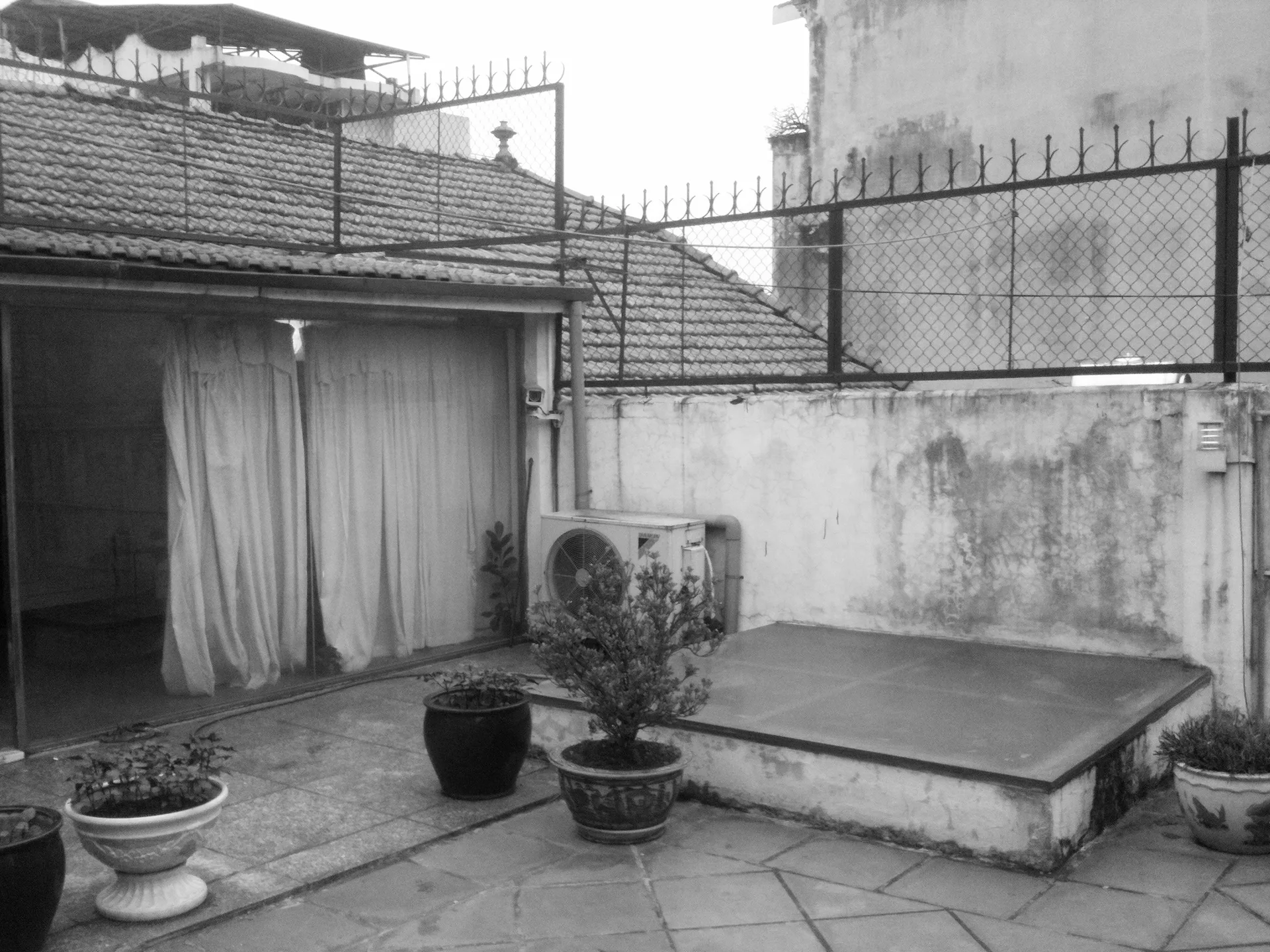 A black and white photo of a weathered courtyard patio featuring irregular stone tile flooring, several potted plants, and a curtained sliding door.