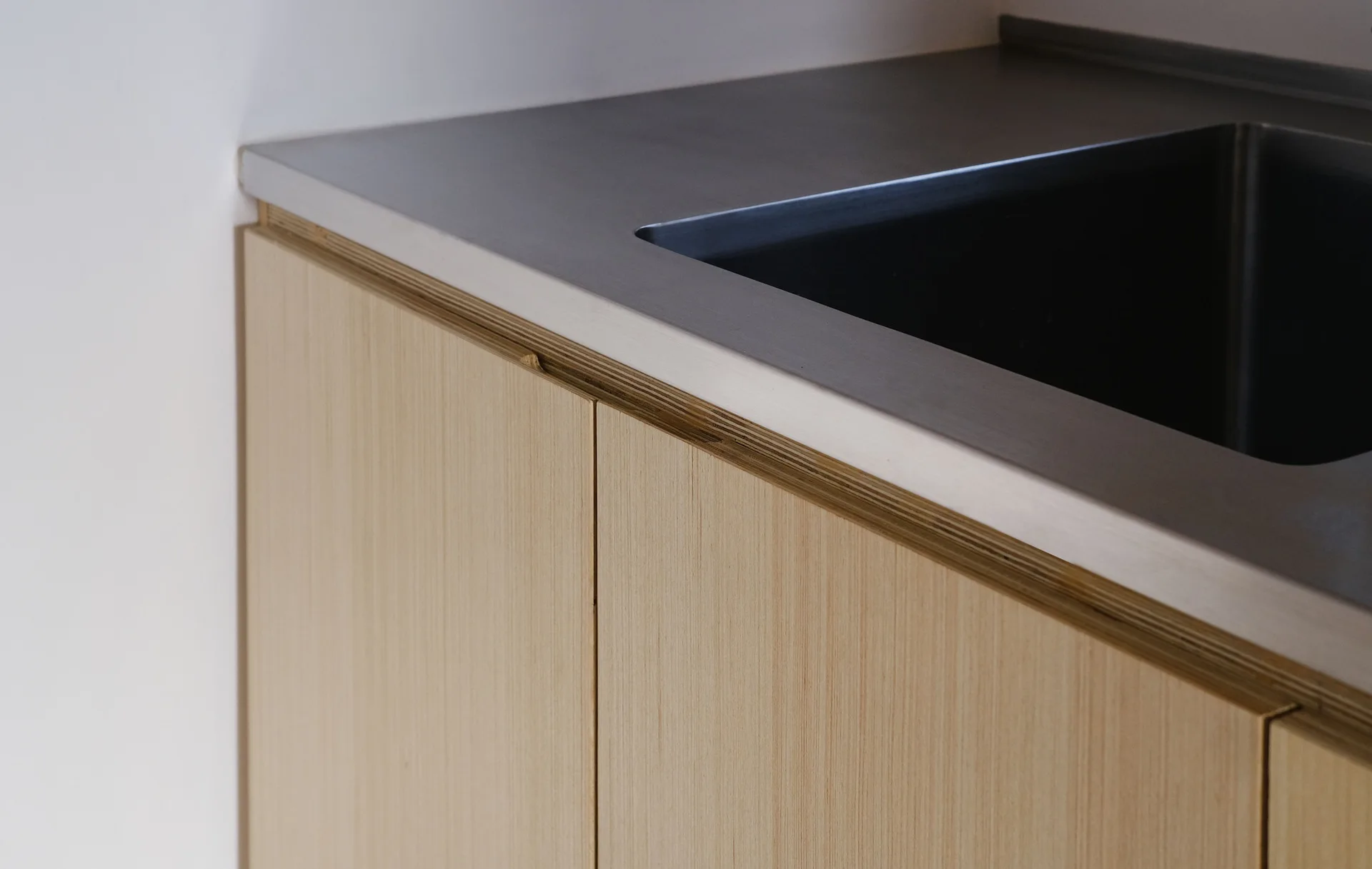 A close-up of light wood kitchen cabinets featuring exposed plywood edges under a matte stainless steel countertop with an integrated sink.