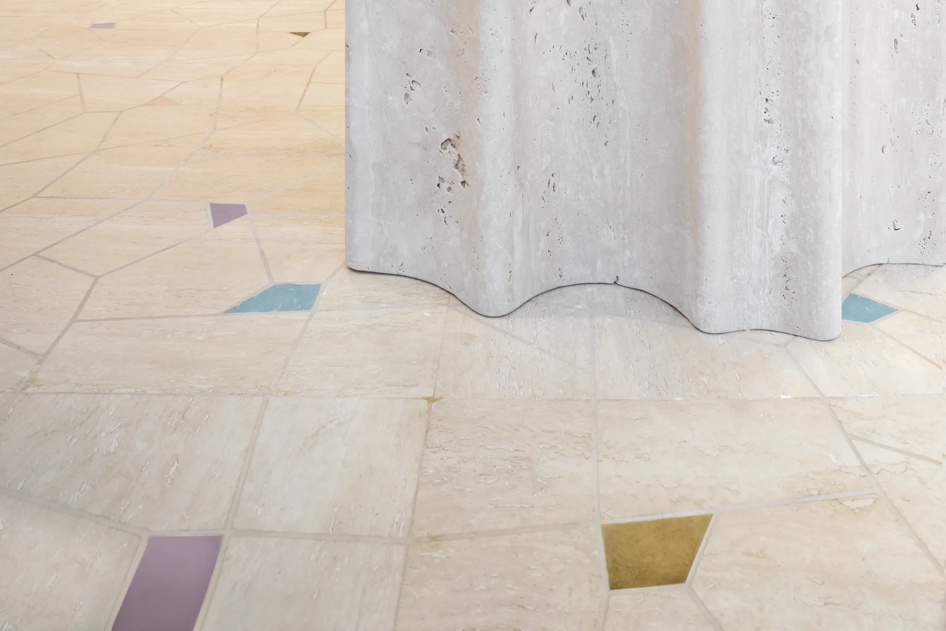 A close-up of a wavy-edged travertine pedestal base resting on beige stone flooring inlaid with small, colorful geometric tile pieces.