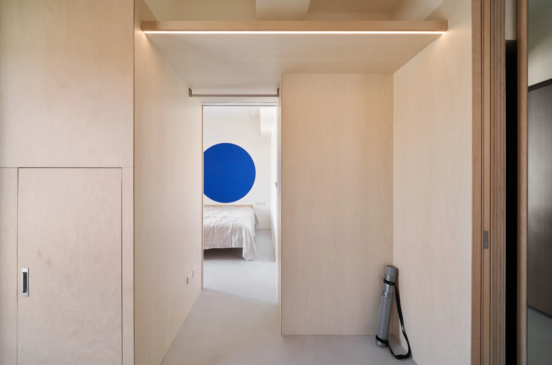 A minimalist hallway lined with light wood paneling and integrated lighting leads to a bedroom featuring a large blue circle on the back wall.