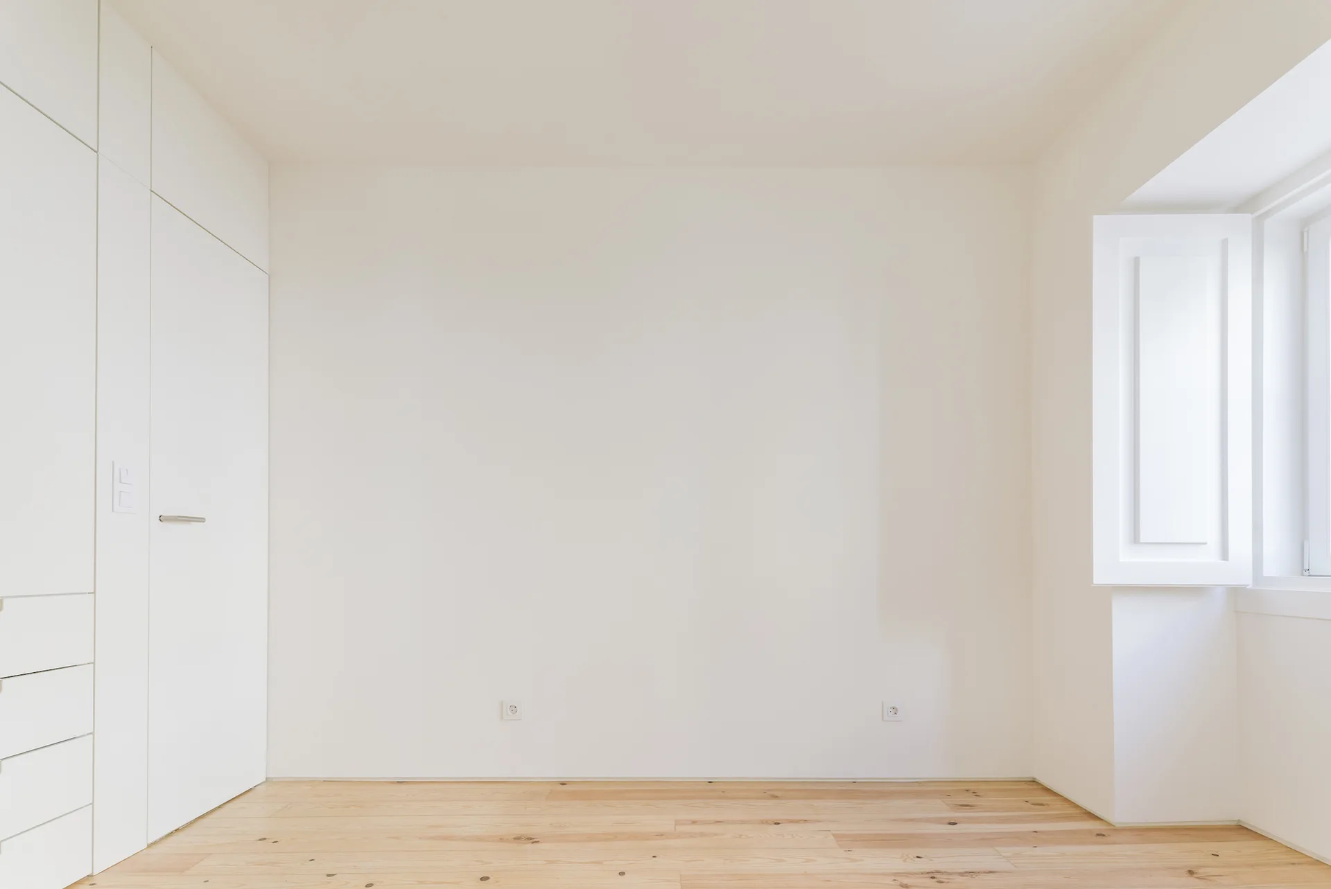 A minimalist empty bedroom features a built-in white wardrobe system, light-colored wood flooring, and a window with white shutters.