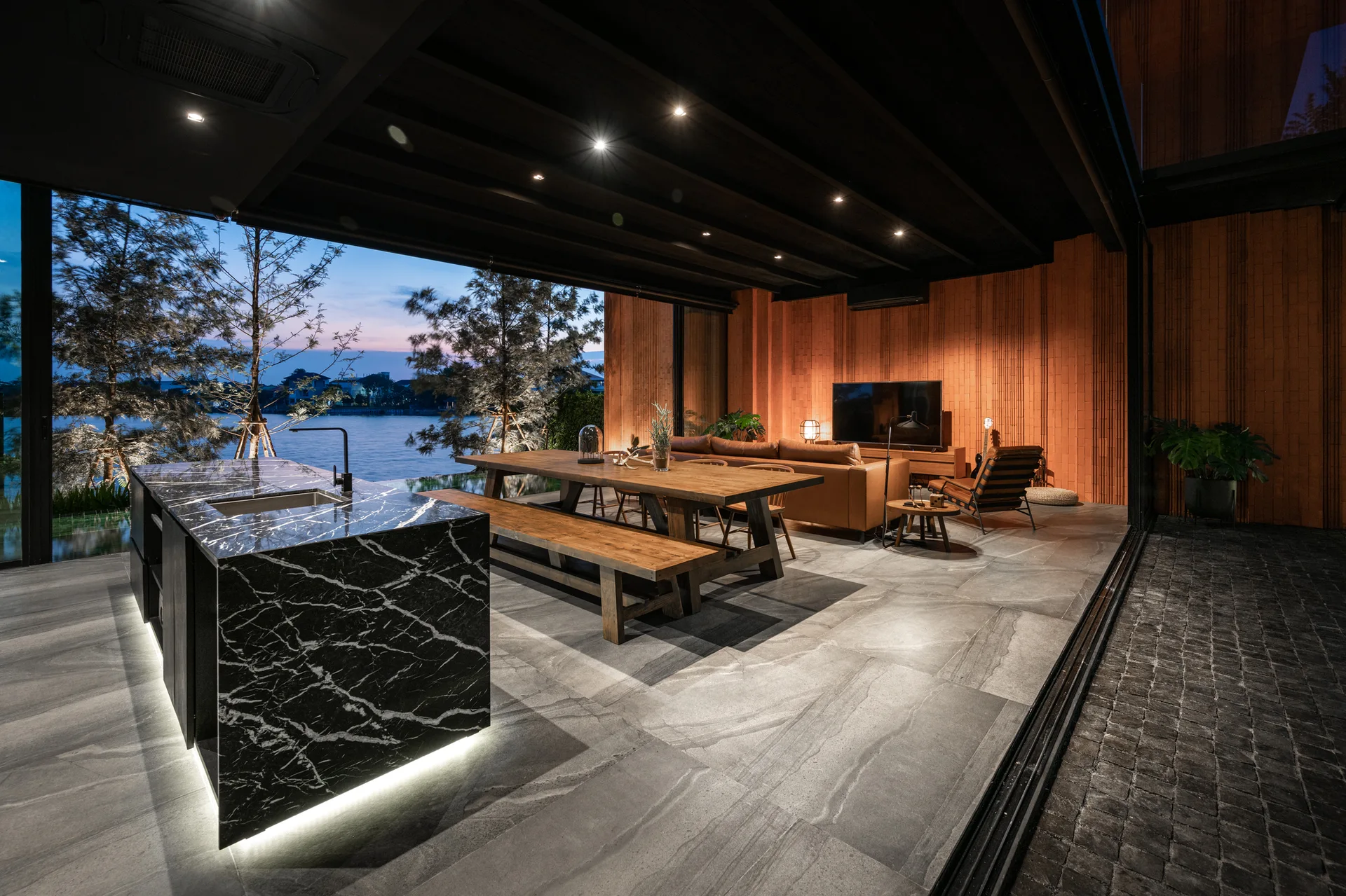 An open-concept room features a black marble kitchen island, a wooden dining table, and a tan sofa next to large sliding glass doors facing a river.
