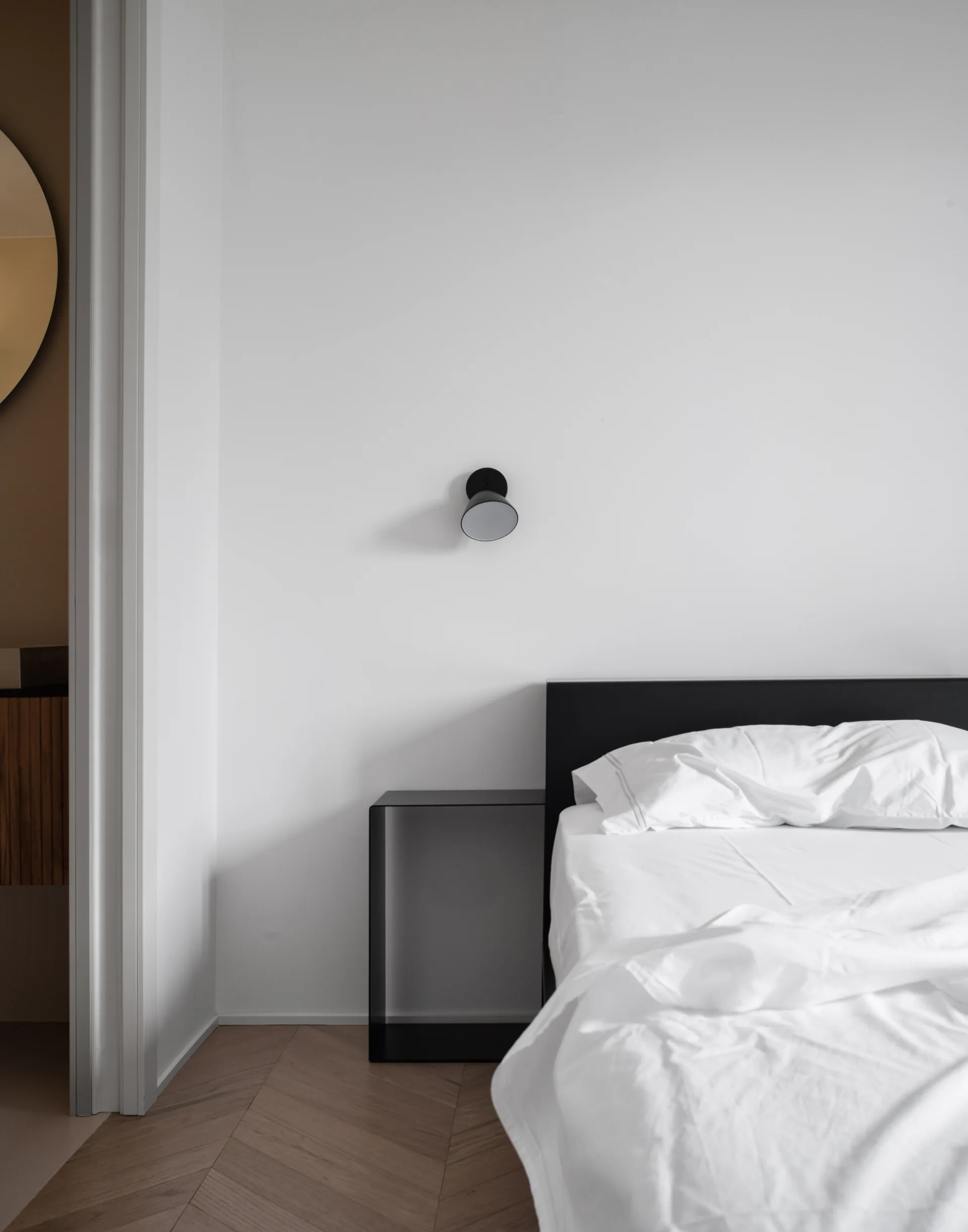 A minimalist bedroom features a bed with white linens and a black headboard next to an open black metal nightstand on herringbone wood floors.