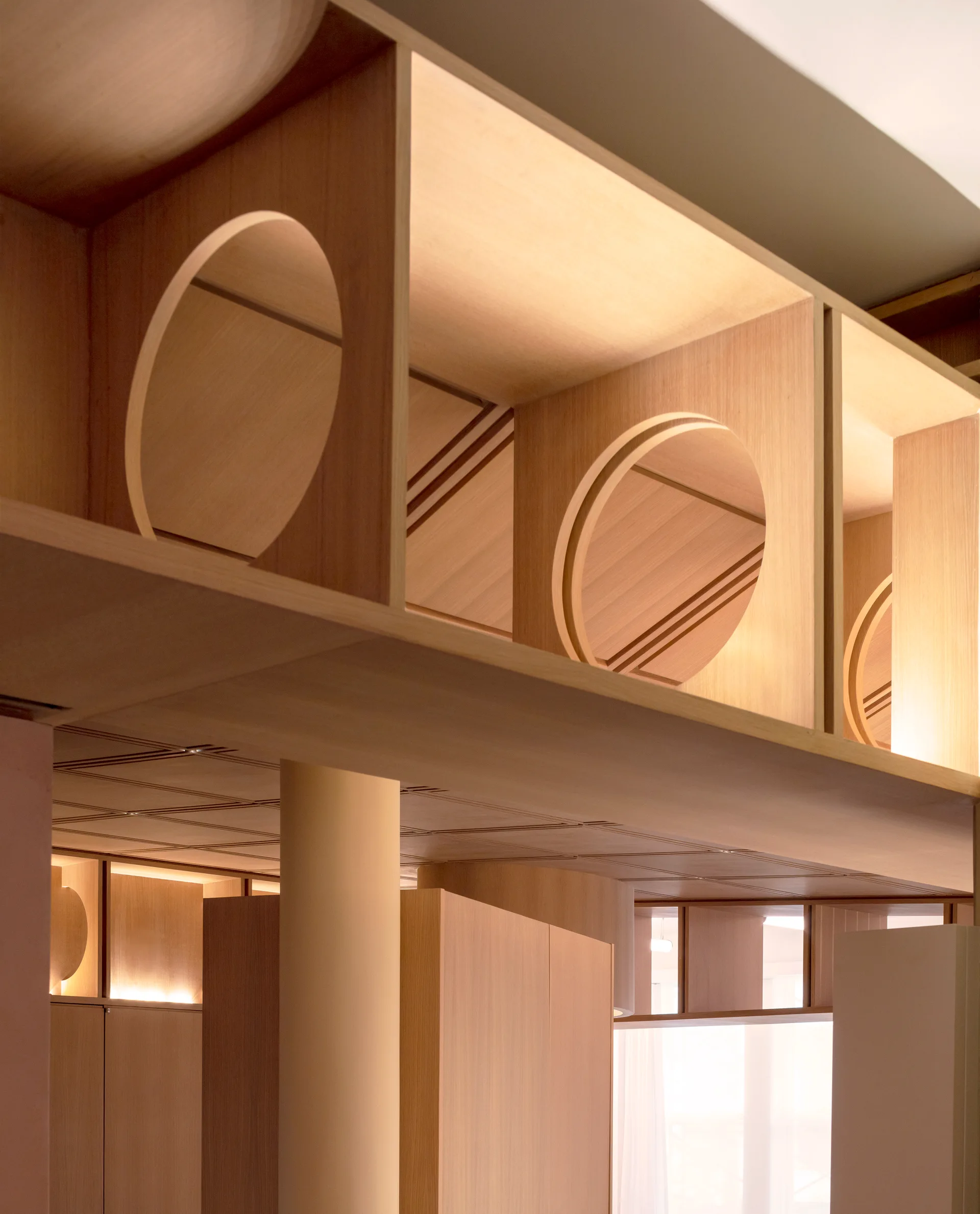 A close-up view of custom light wood millwork featuring geometric circular cutouts, integrated shelf lighting, and a cylindrical support column below.