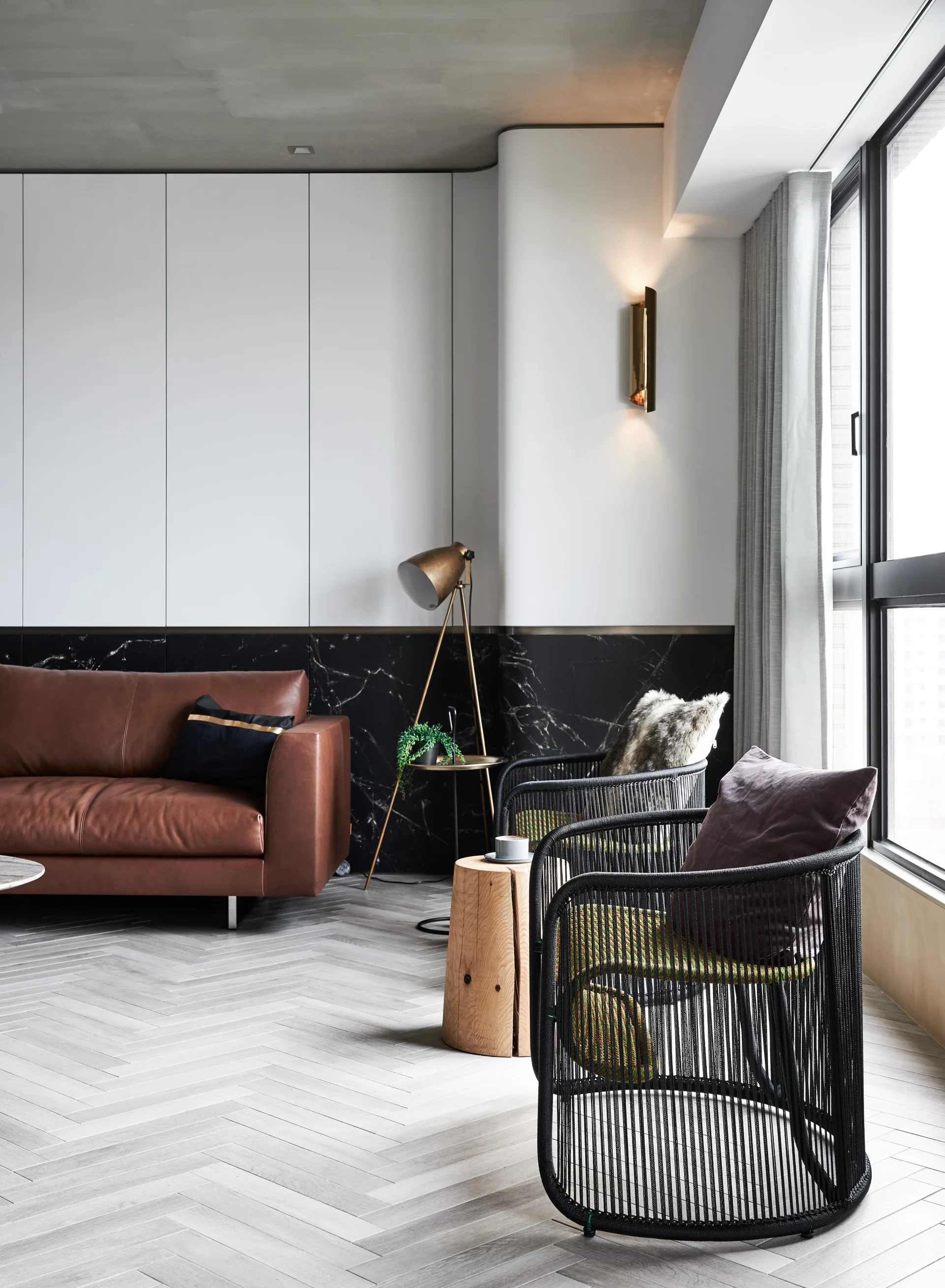 A modern living room features a brown leather sofa, black woven armchairs, and a log side table on light grey herringbone flooring.