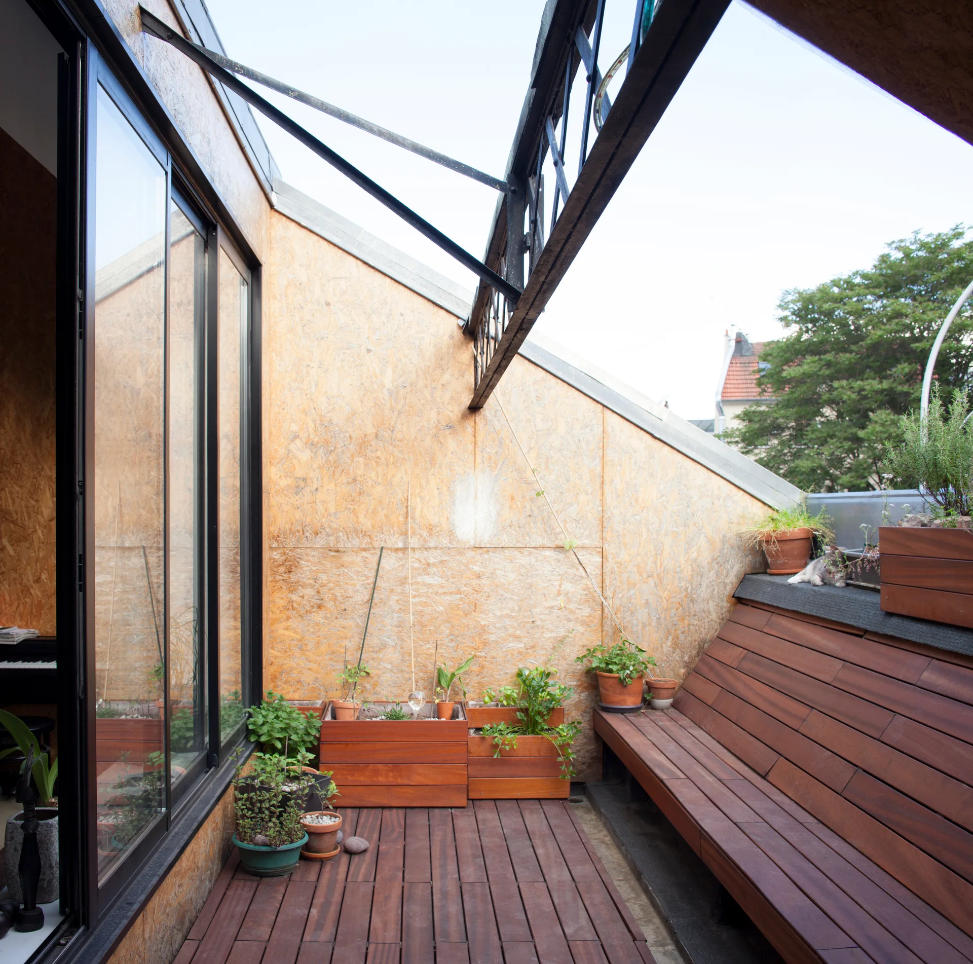 An outdoor terrace features OSB paneled walls, a built-in wooden bench, wood plank decking, and small potted plants.
