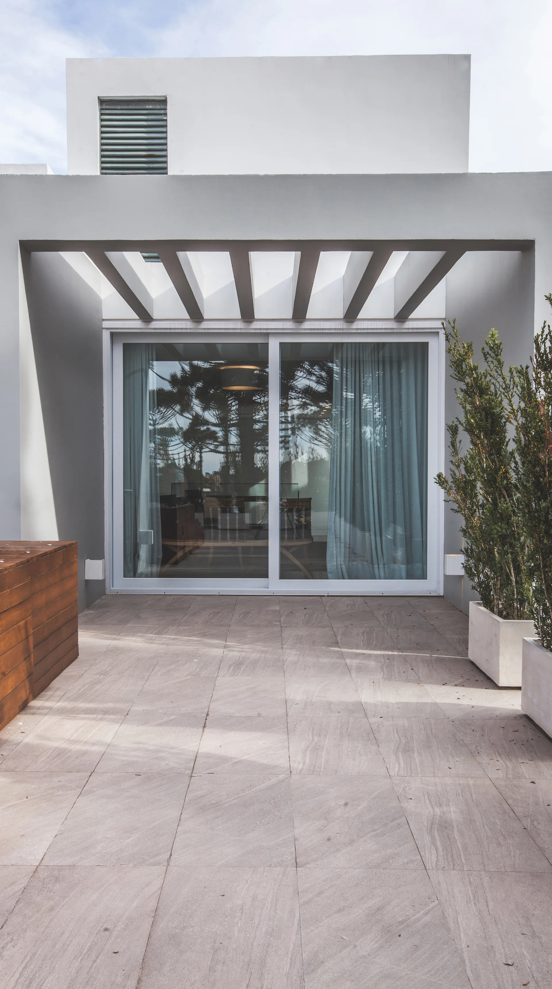 A minimalist stone tile patio with large sliding glass doors, a concrete beam pergola overhead, and white rectangular planters.