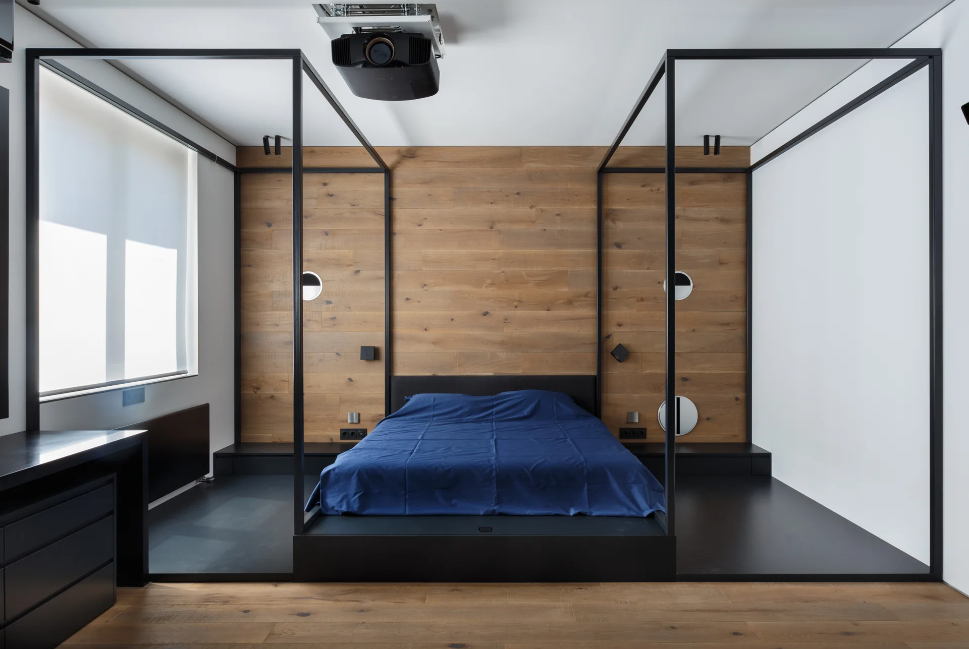 A minimalist bedroom featuring a black platform bed with blue sheets and a metal canopy frame against a horizontal wood-paneled accent wall.
