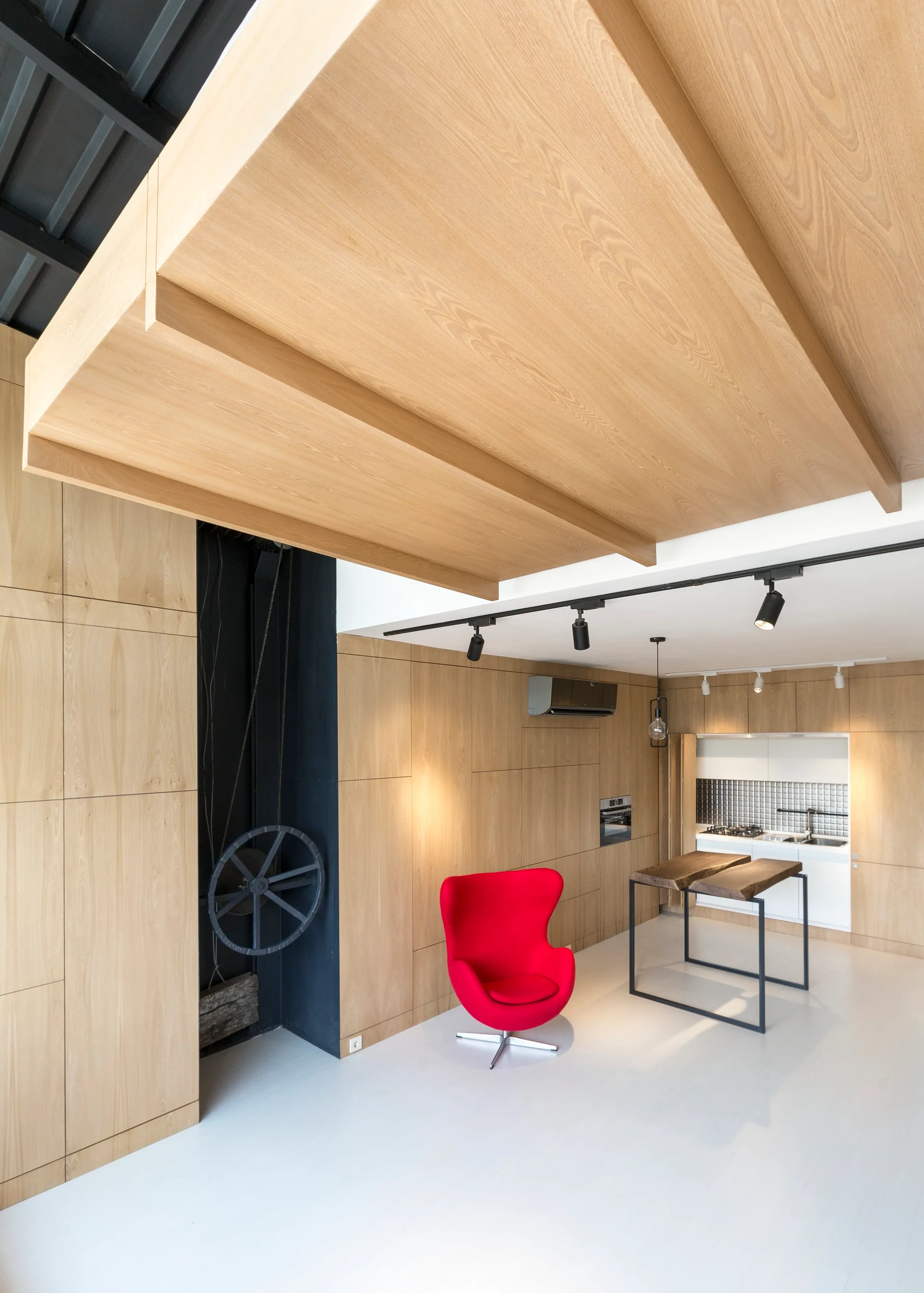 A modern kitchen space features light wood paneled walls, a large wood ceiling panel, a bright red armchair, and a metal-framed island.