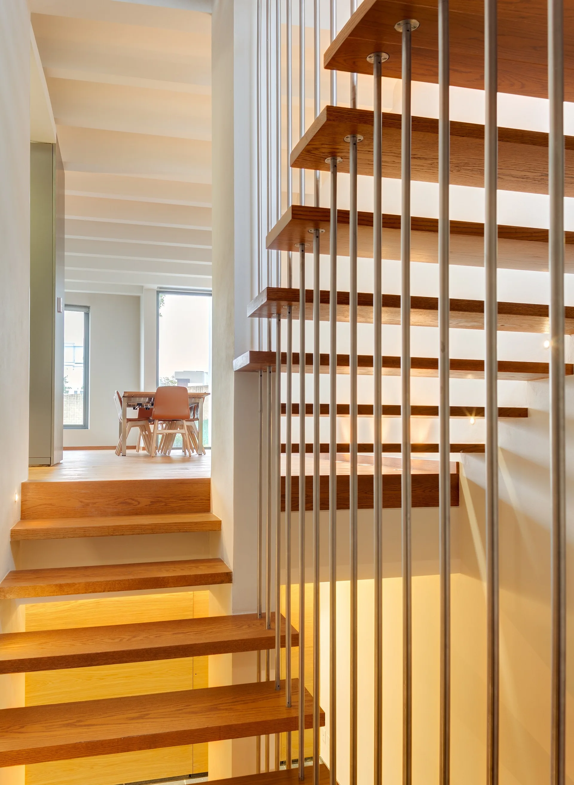 A modern open-riser wood staircase with thin vertical metal railings and integrated under-tread lighting, showing a dining area in the background.