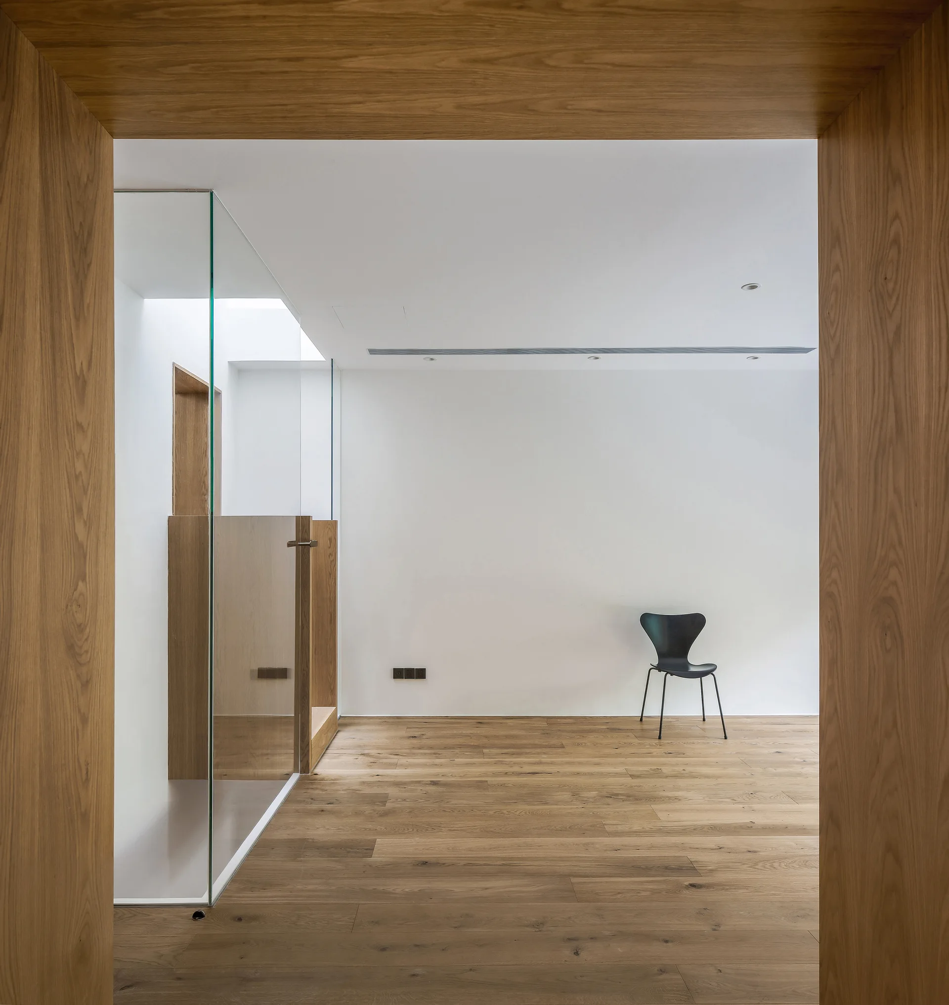 A view through a thick wood-paneled doorway into a minimalist white room with wood flooring, a clear glass partition, and a single black chair.