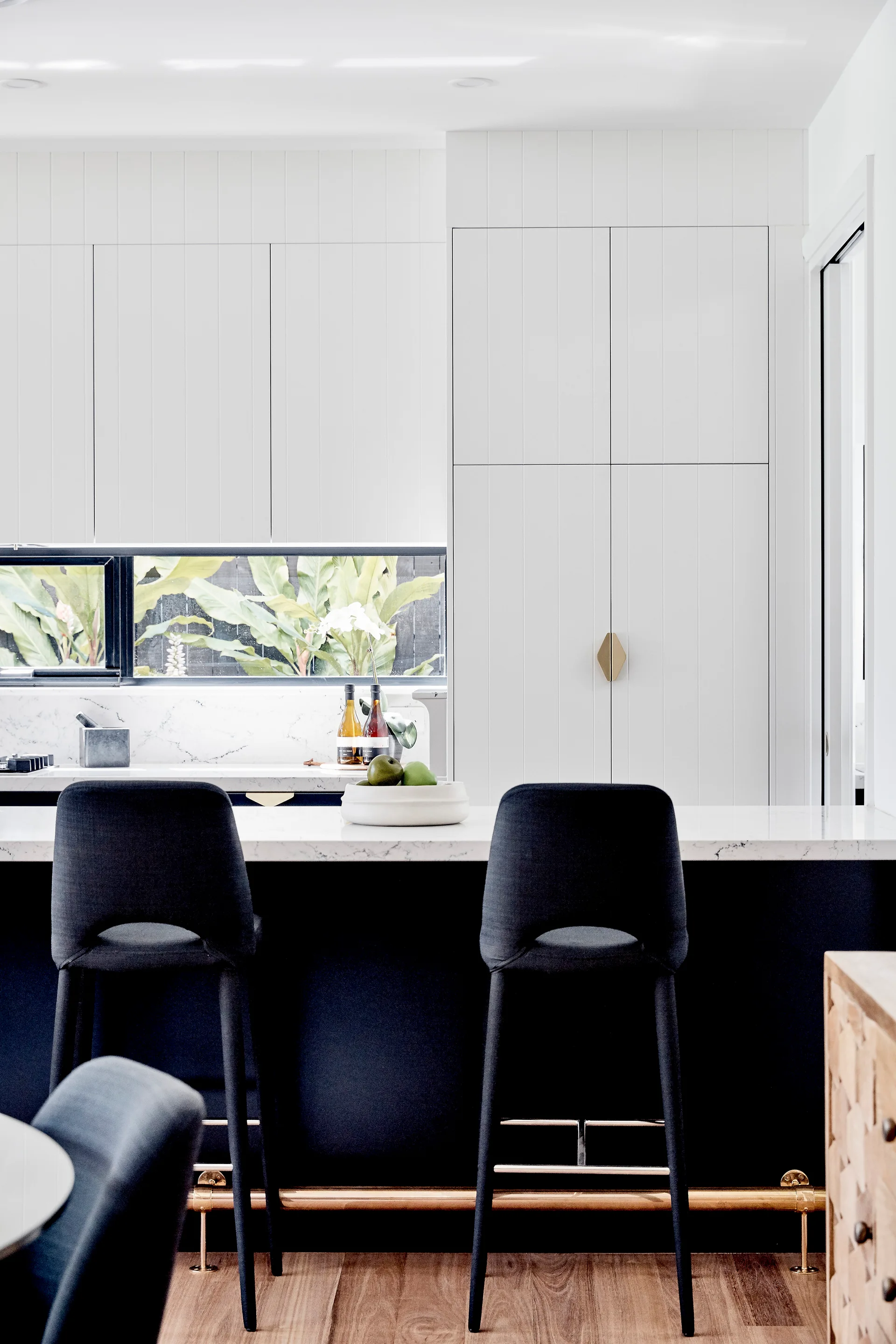 A modern kitchen features a black island with a marble countertop, two dark bar stools, and white vertical paneled cabinets beneath a horizontal window.