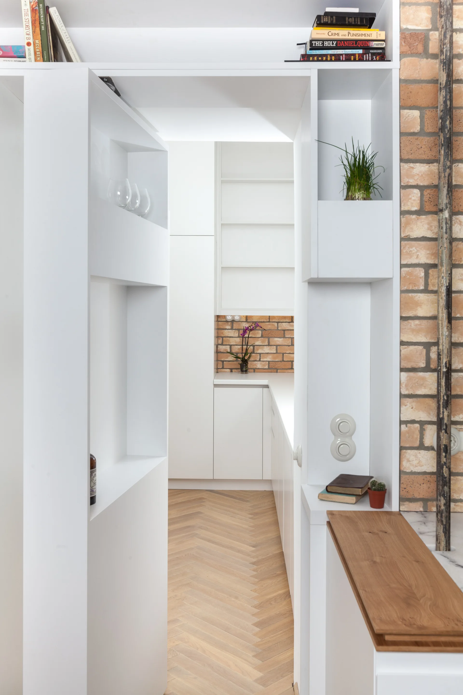 White built-in cabinetry frames a doorway leading to a kitchen with herringbone wood floors and an exposed brick accent wall.