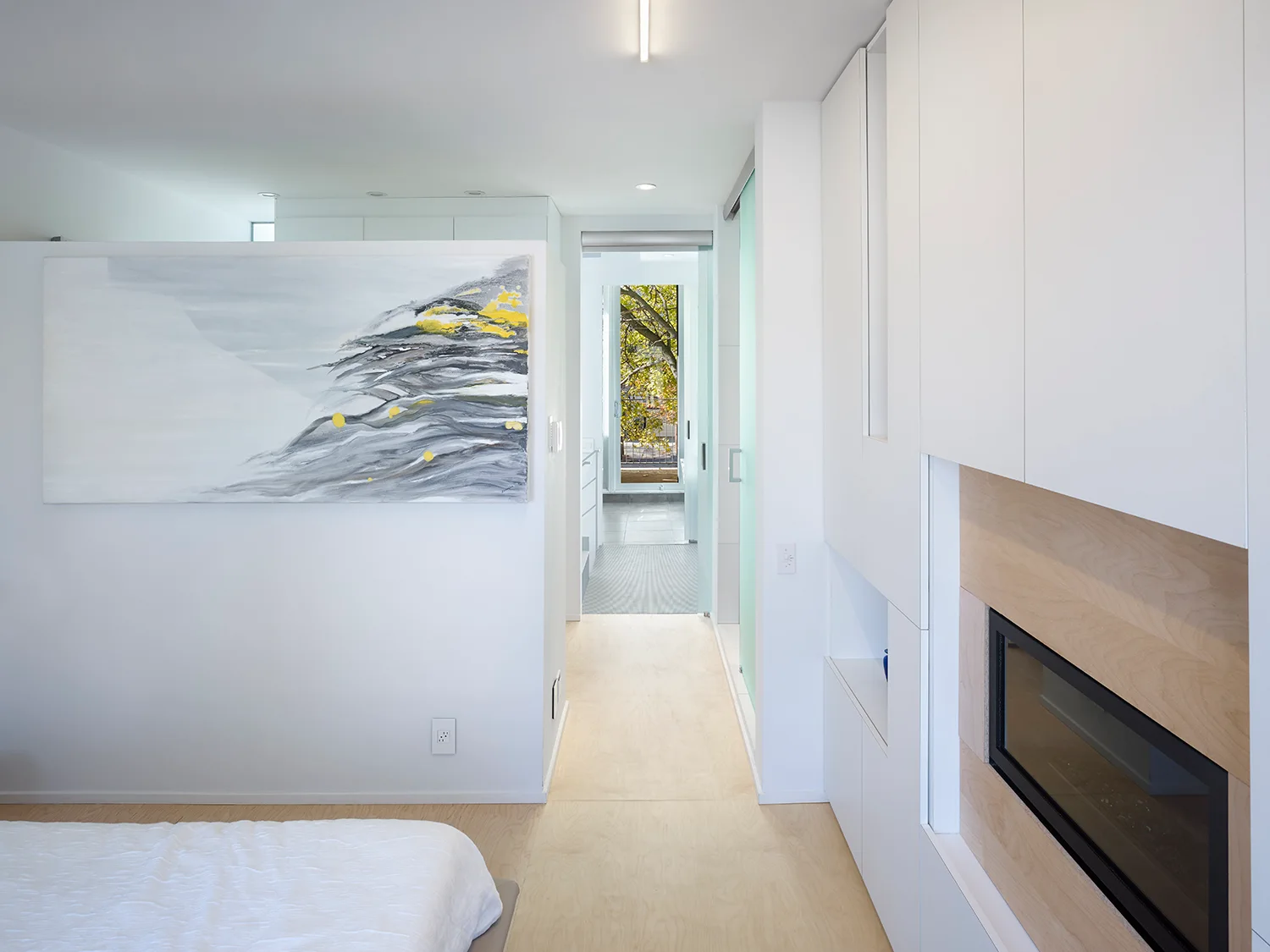 A minimalist white bedroom hallway featuring light wood flooring, a large abstract painting, and white built-in storage with an integrated fireplace.