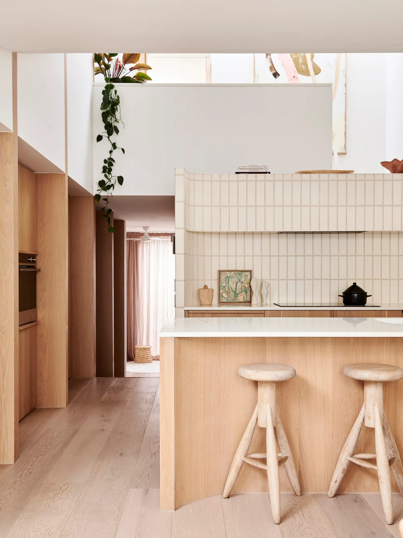 A minimalist kitchen featuring a light oak island with wooden stools, a cream-colored vertical tile backsplash, and light wood flooring.