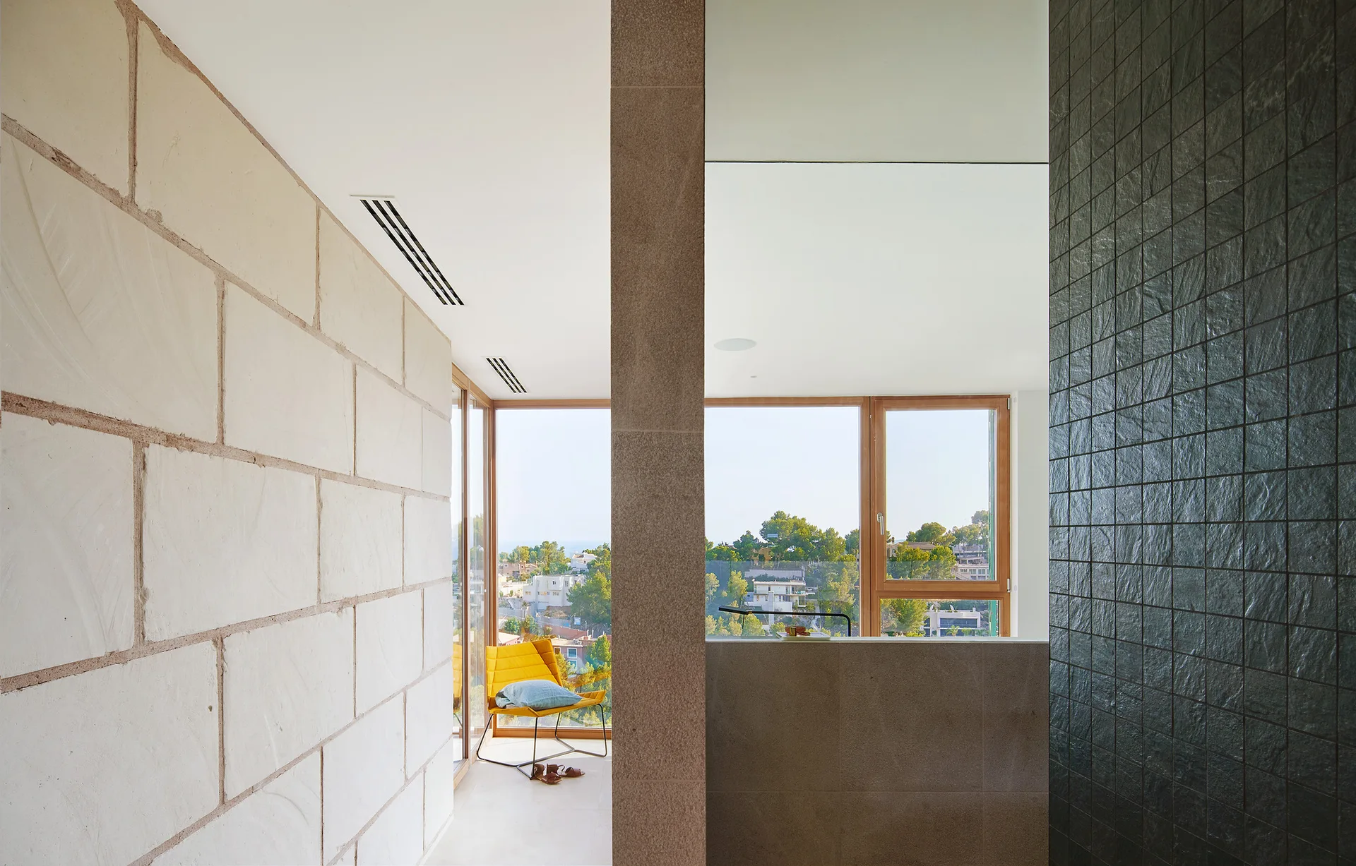 A modern room features a large textured stone wall, a central pillar, a dark tiled wall, and a yellow armchair near floor-to-ceiling windows.