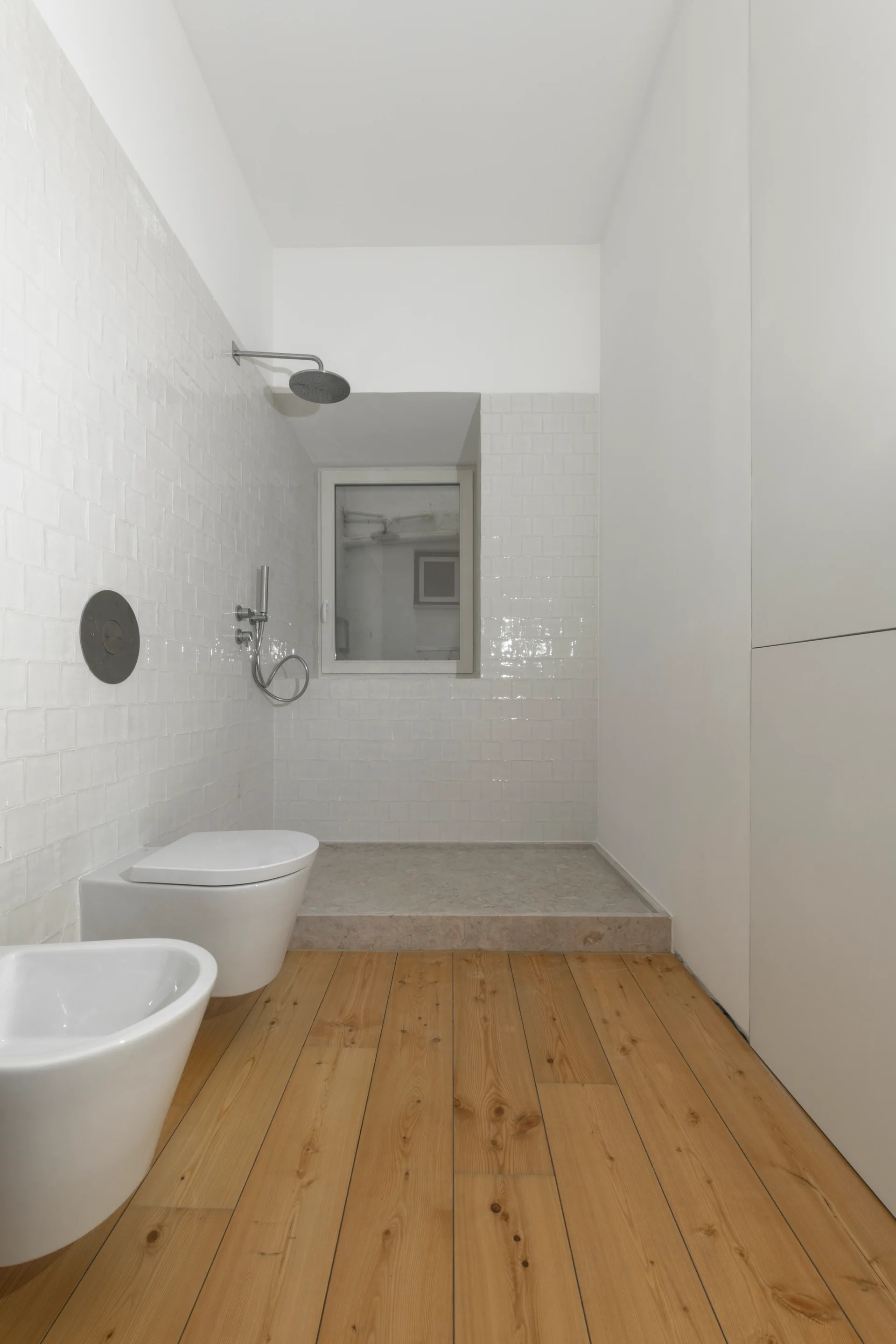 Minimalist white tiled bathroom featuring a walk-in shower, wall-mounted toilet and bidet, and natural wood plank flooring.