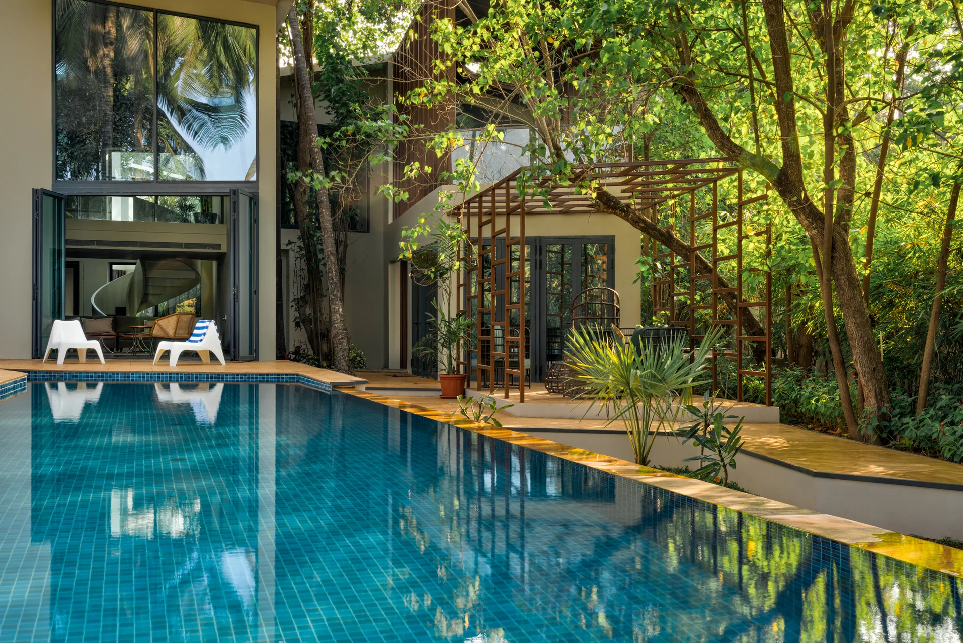 A backyard swimming pool with blue tiles sits next to a modern house with folding glass doors and a metal pergola under tall trees.