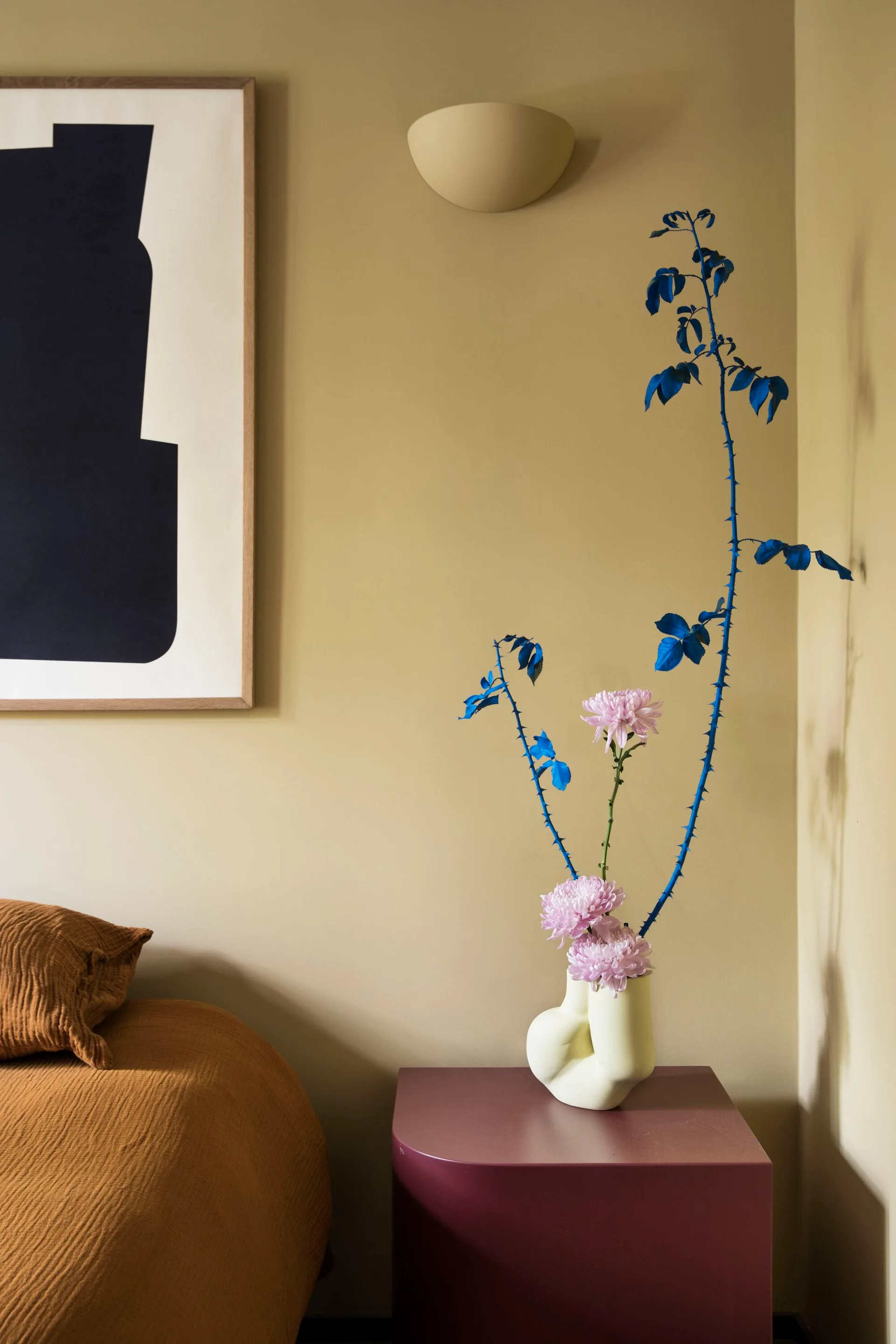 A minimalist bedroom corner features a plum-colored bedside table with a sculptural white vase and a framed abstract print against a muted olive wall.