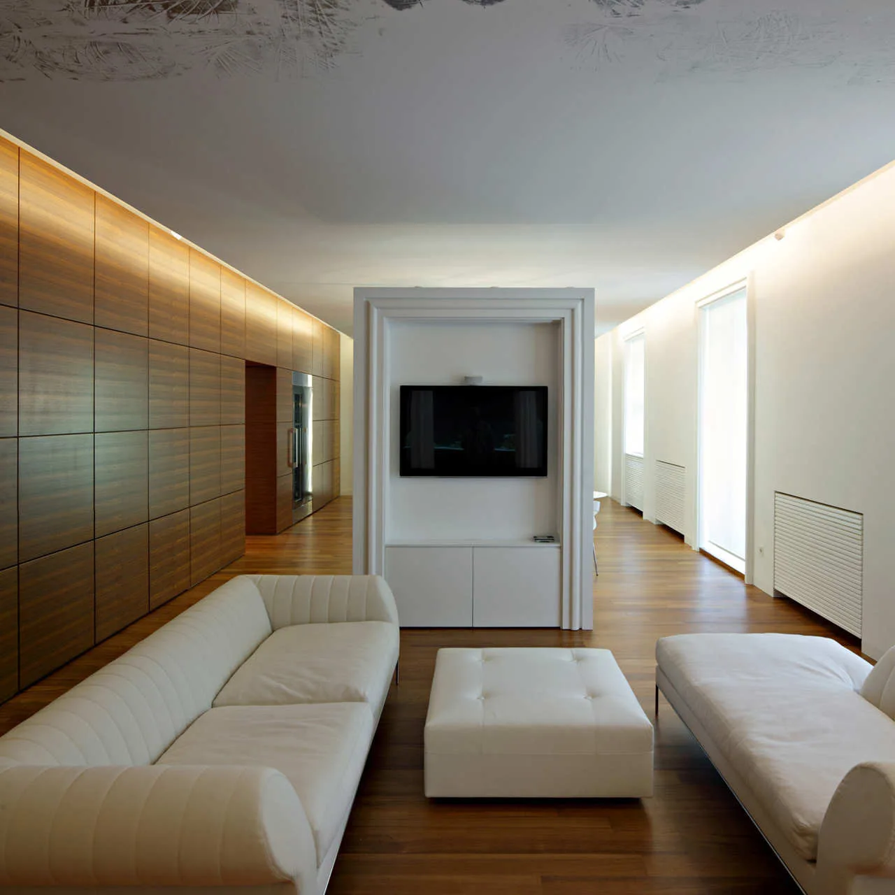 A minimalist living room with white sofas, a central white partition wall mounting a TV, and a warm wood-paneled wall.