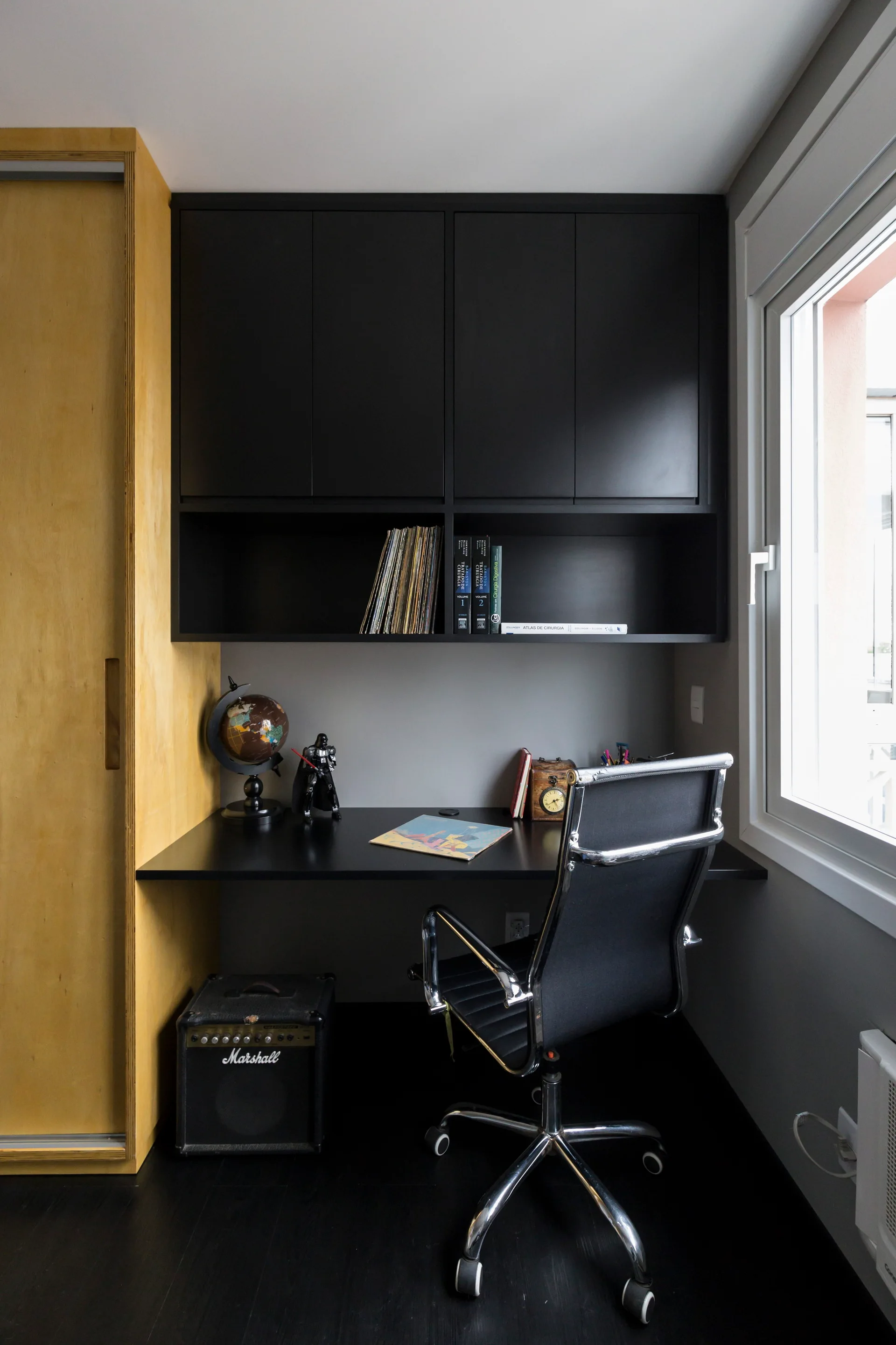 A minimalist home office nook featuring a matte black built-in desk and cabinets beside a light wood wardrobe and a black leather rolling chair.