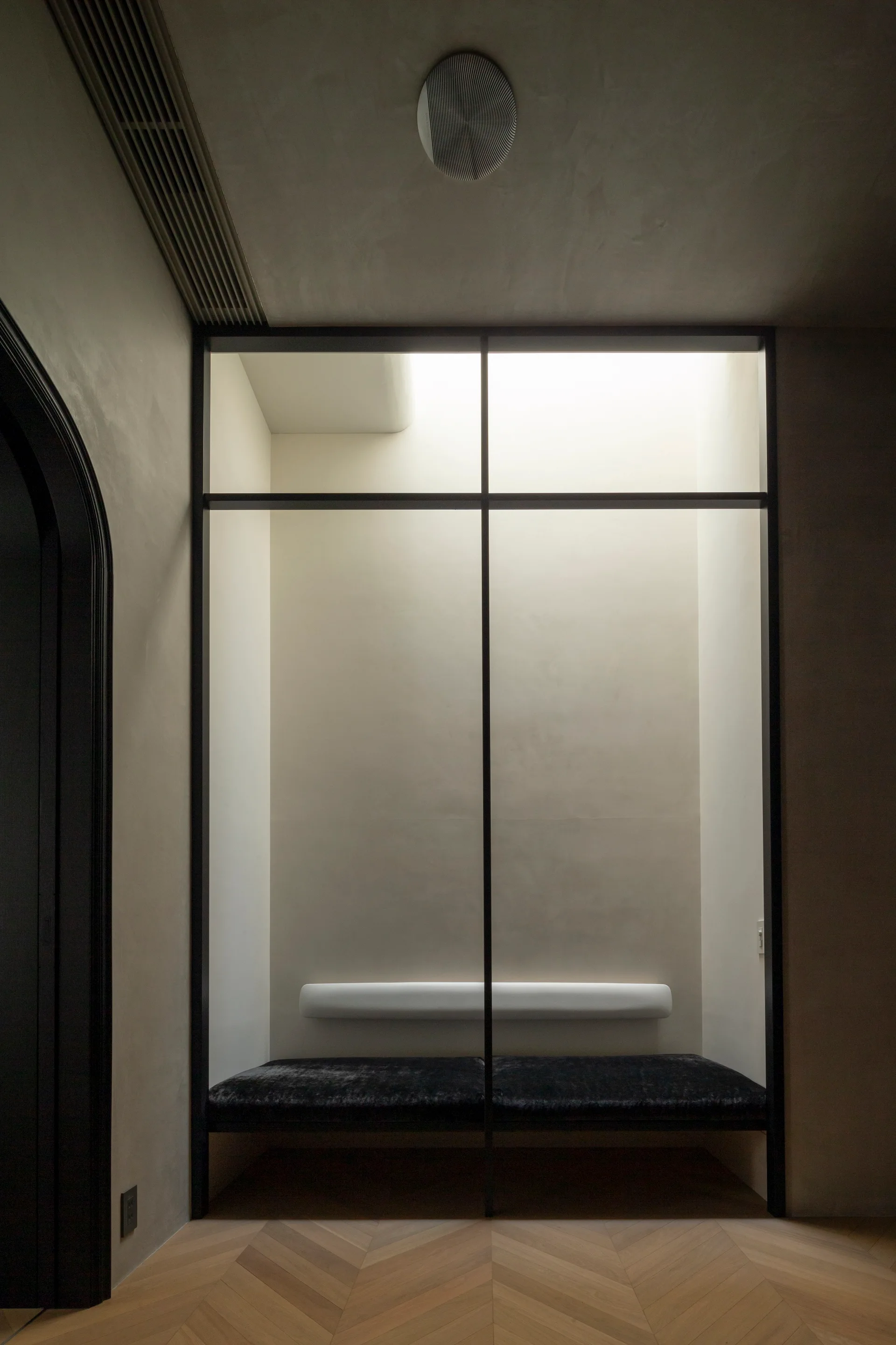 A minimalist hallway alcove features a built-in dark velvet bench, a cylindrical white bolster backrest, a black-framed glass partition, and a skylight.