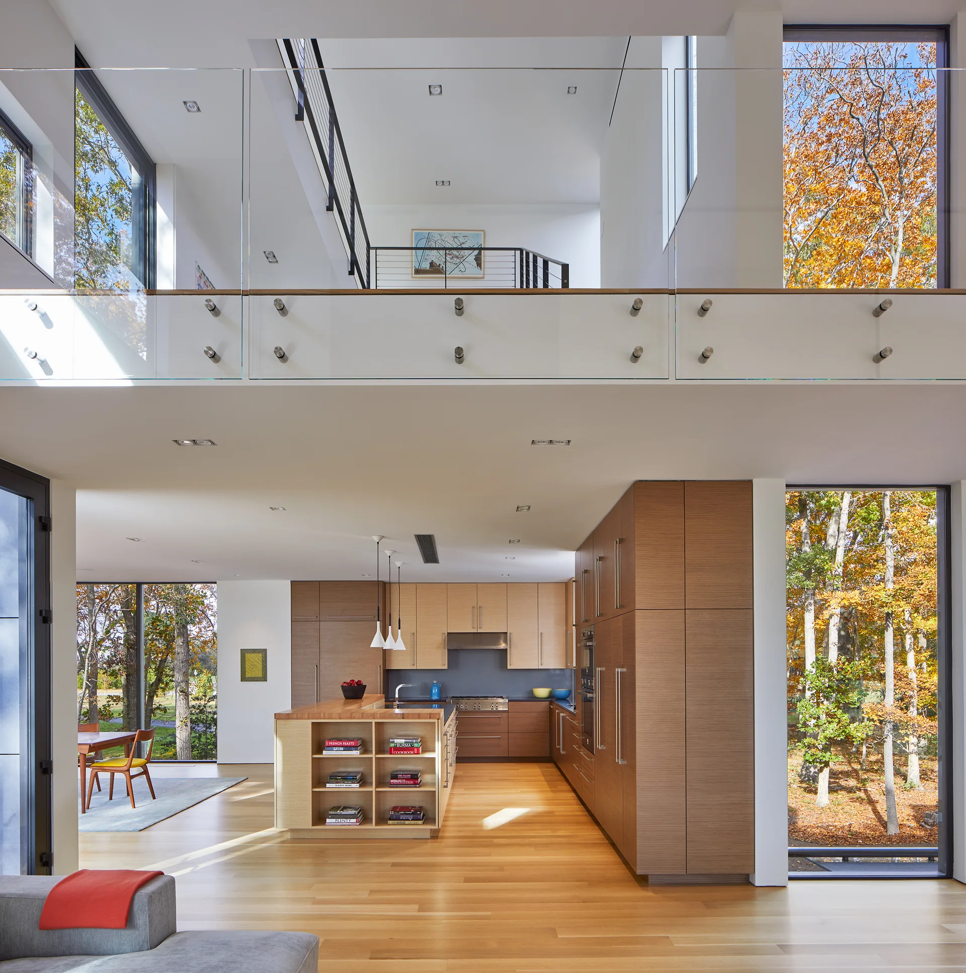 A modern open-plan kitchen features wood cabinetry, a central island with built-in shelving, and an overlooking mezzanine level with a glass railing.