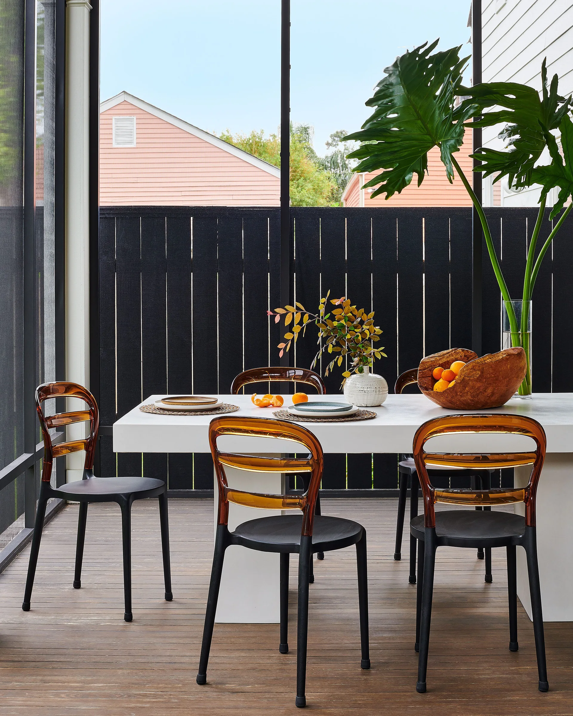 A modern white dining table paired with translucent amber and black chairs sits on a wood-decked screened-in porch.
