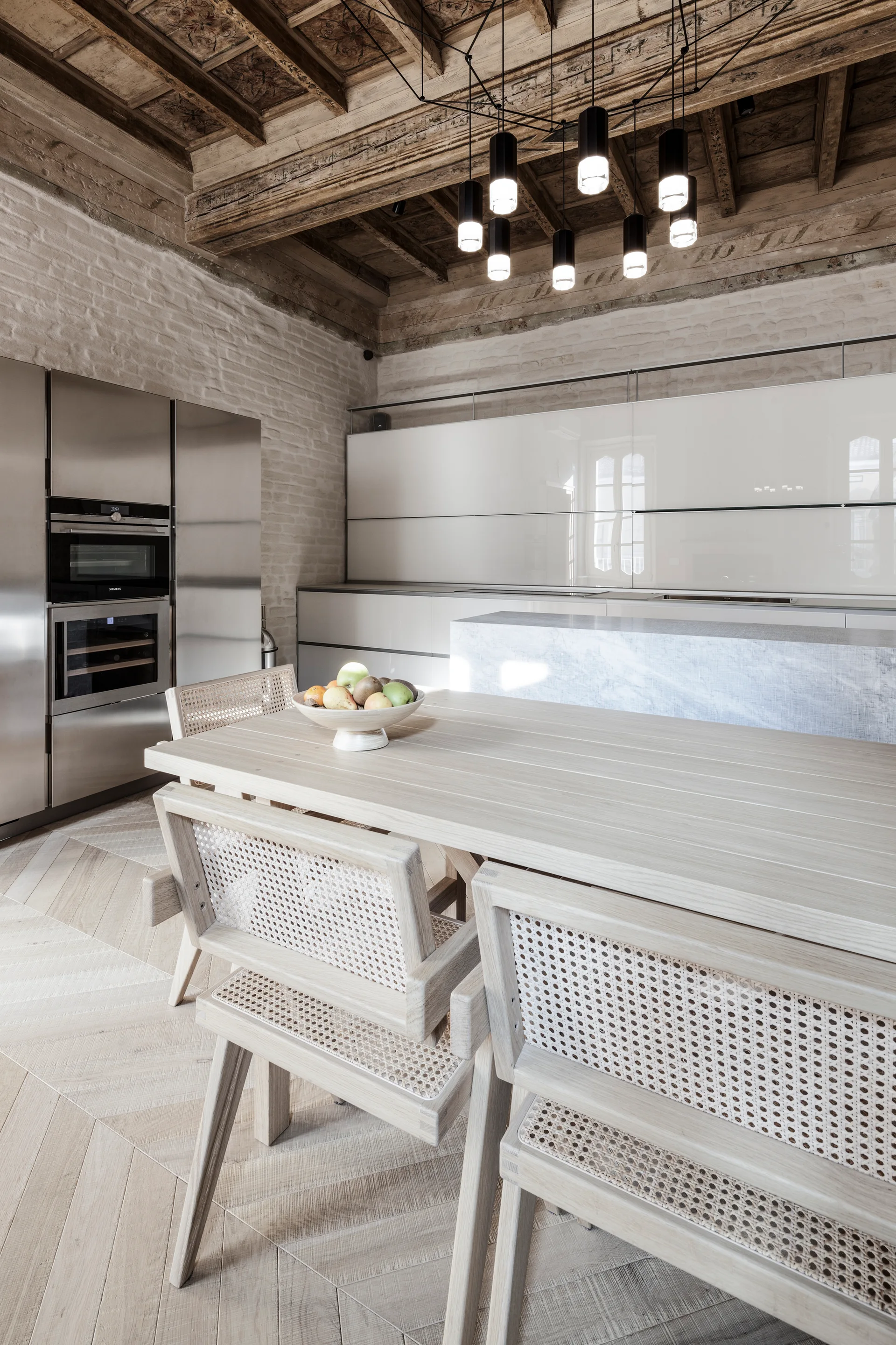 A modern kitchen and dining space features a light wood table with cane back chairs, an exposed beam ceiling, and sleek cabinetry.