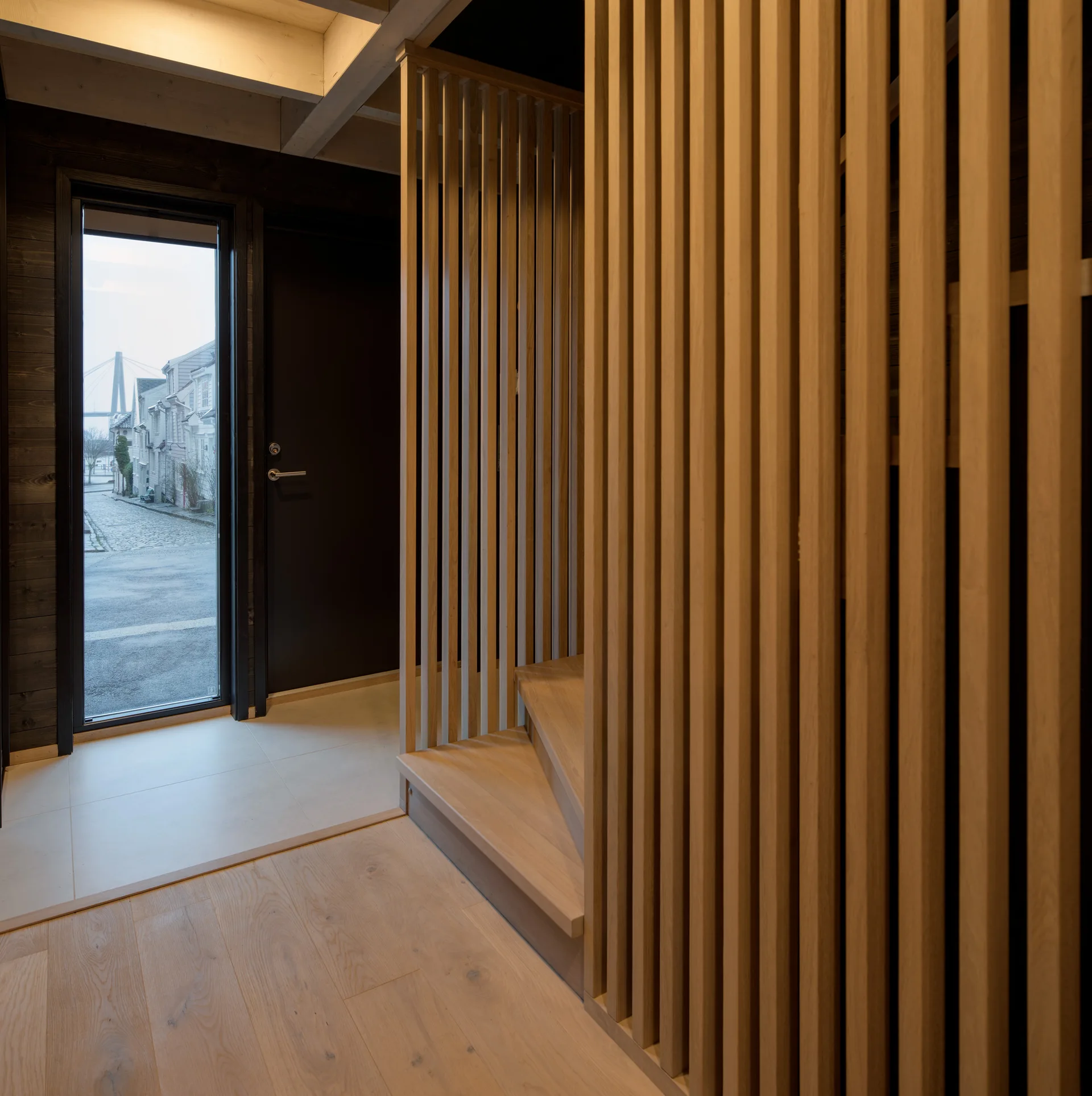 A modern entryway features a vertical light wood slat partition beside a staircase, with a glass door looking out onto a narrow street.