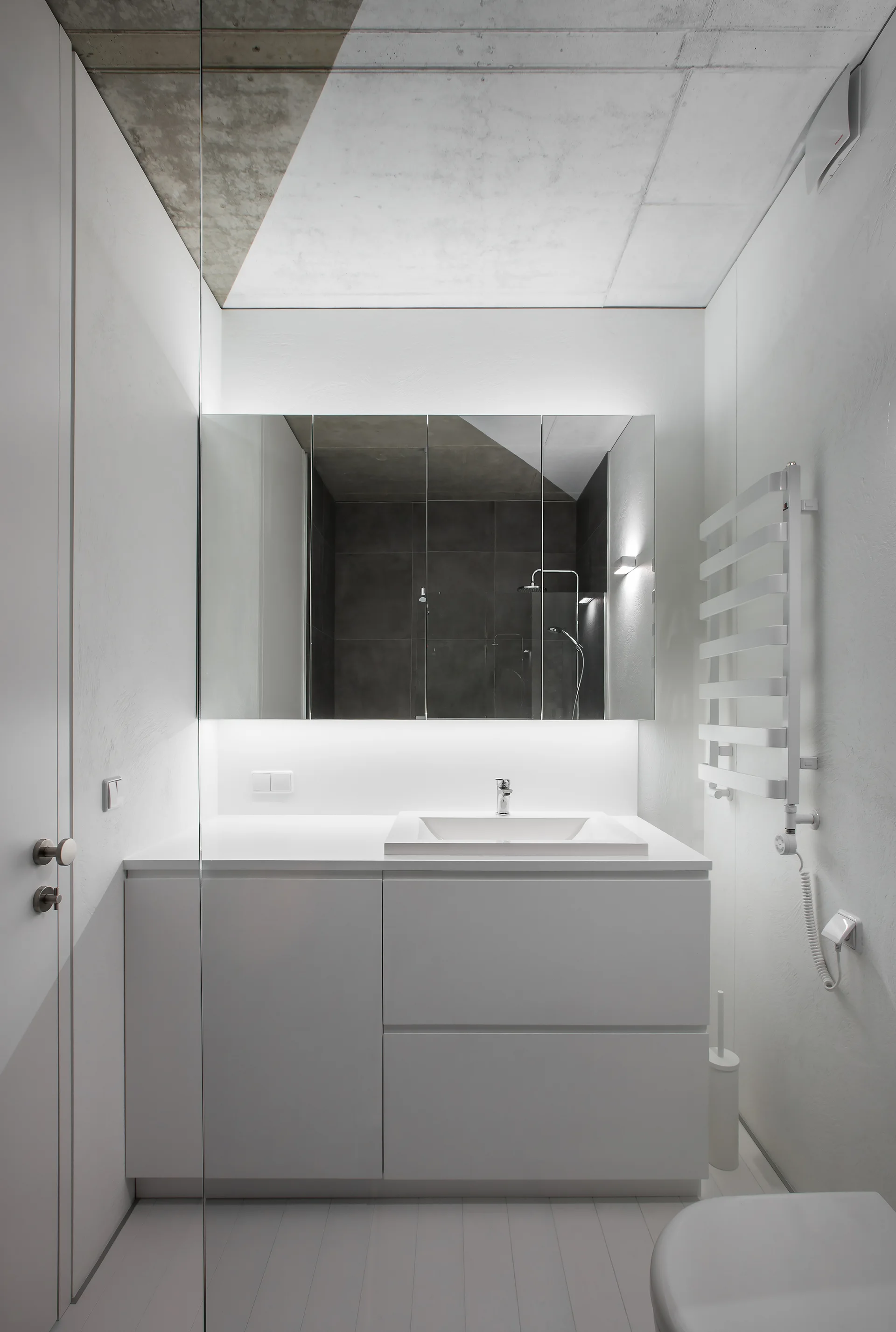 A minimalist white bathroom featuring a floating vanity, a large backlit mirror, and an exposed raw concrete ceiling.