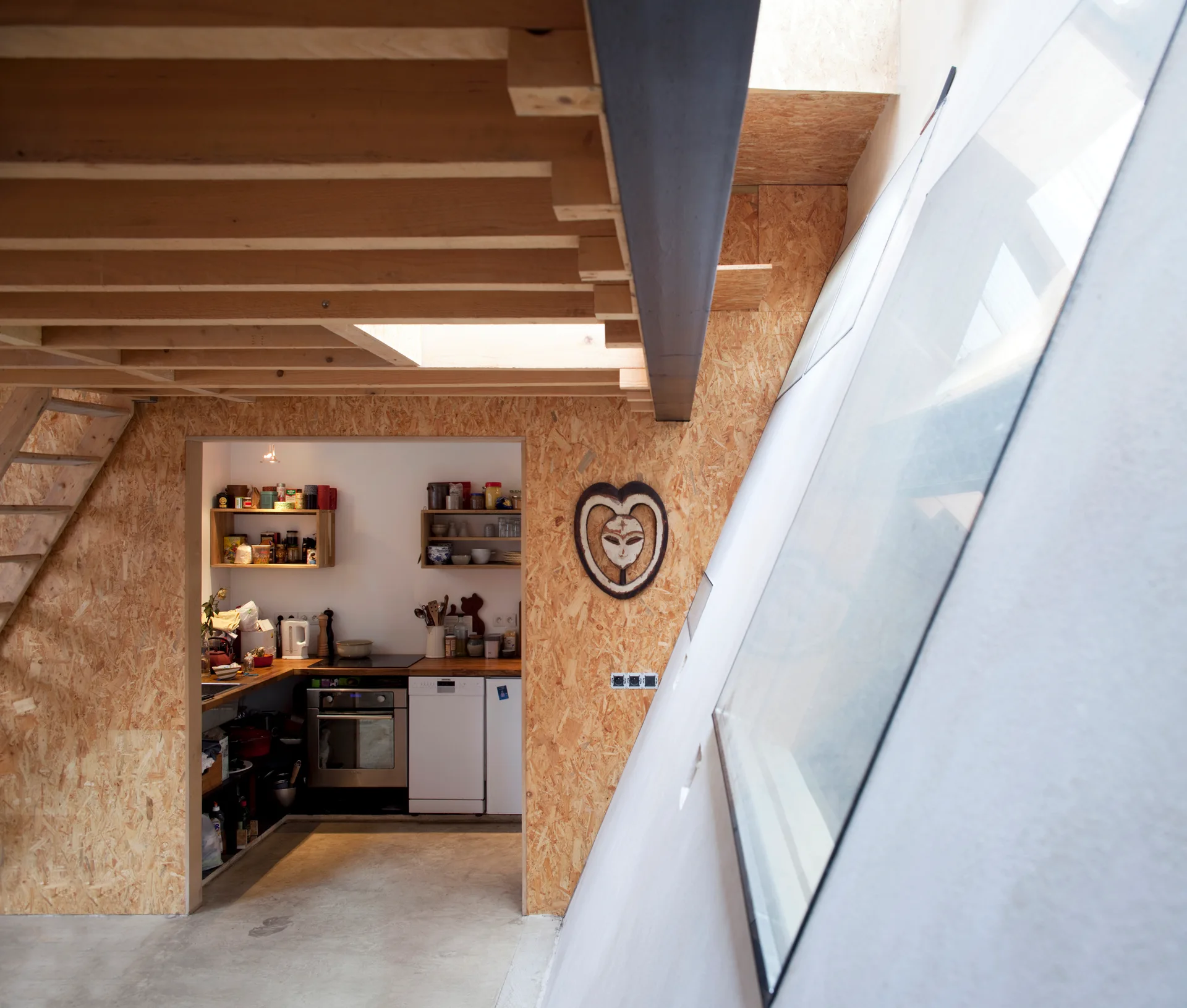 An industrial loft features OSB paneled walls, exposed ceiling joists, a concrete floor, and a sloped skylight looking into a kitchen nook.