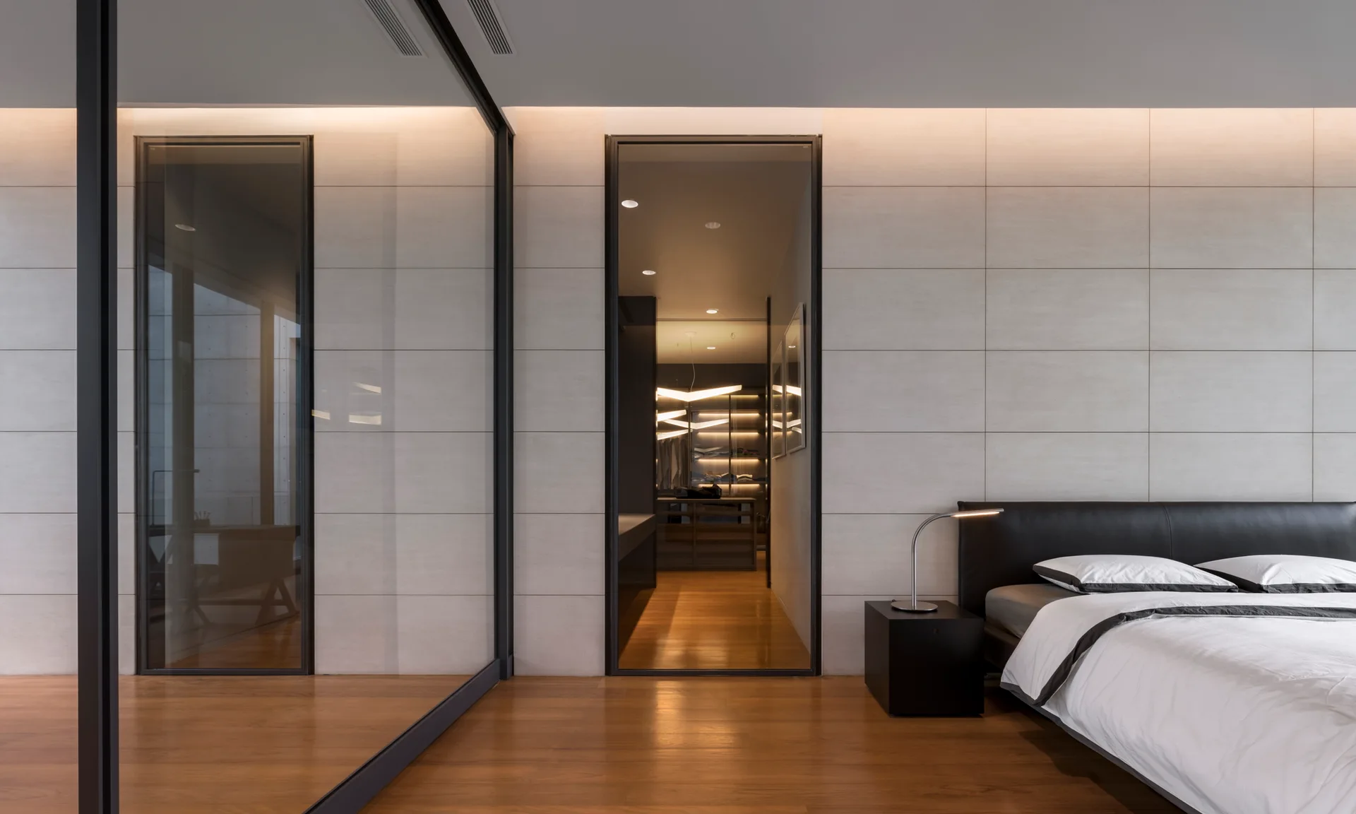 A modern bedroom features a black leather headboard, a tiled accent wall with cove lighting, and a glass partition looking into a closet.