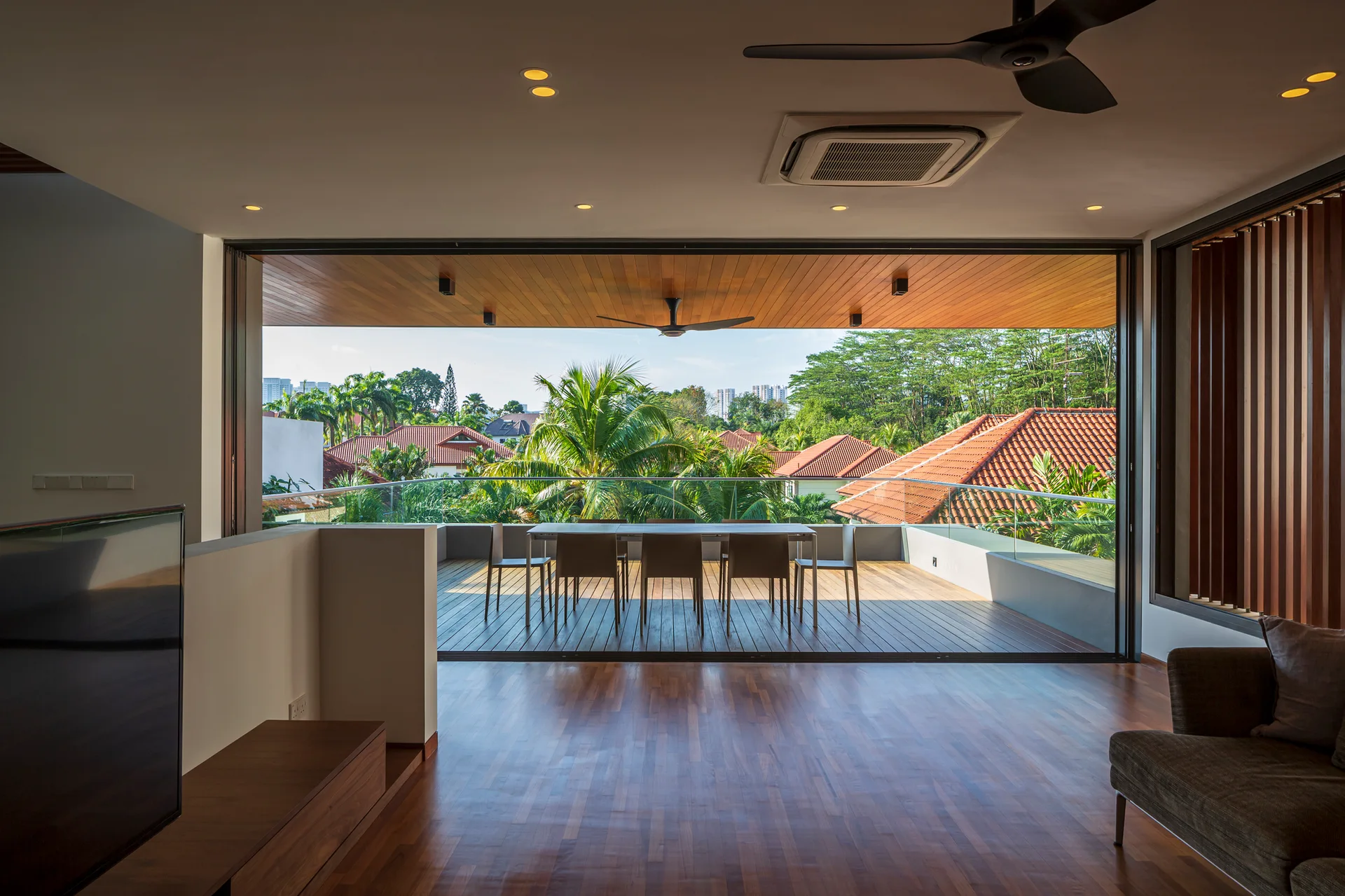 A modern living room opens completely to a spacious wooden deck balcony with an outdoor dining table and glass railing overlooking tropical trees.