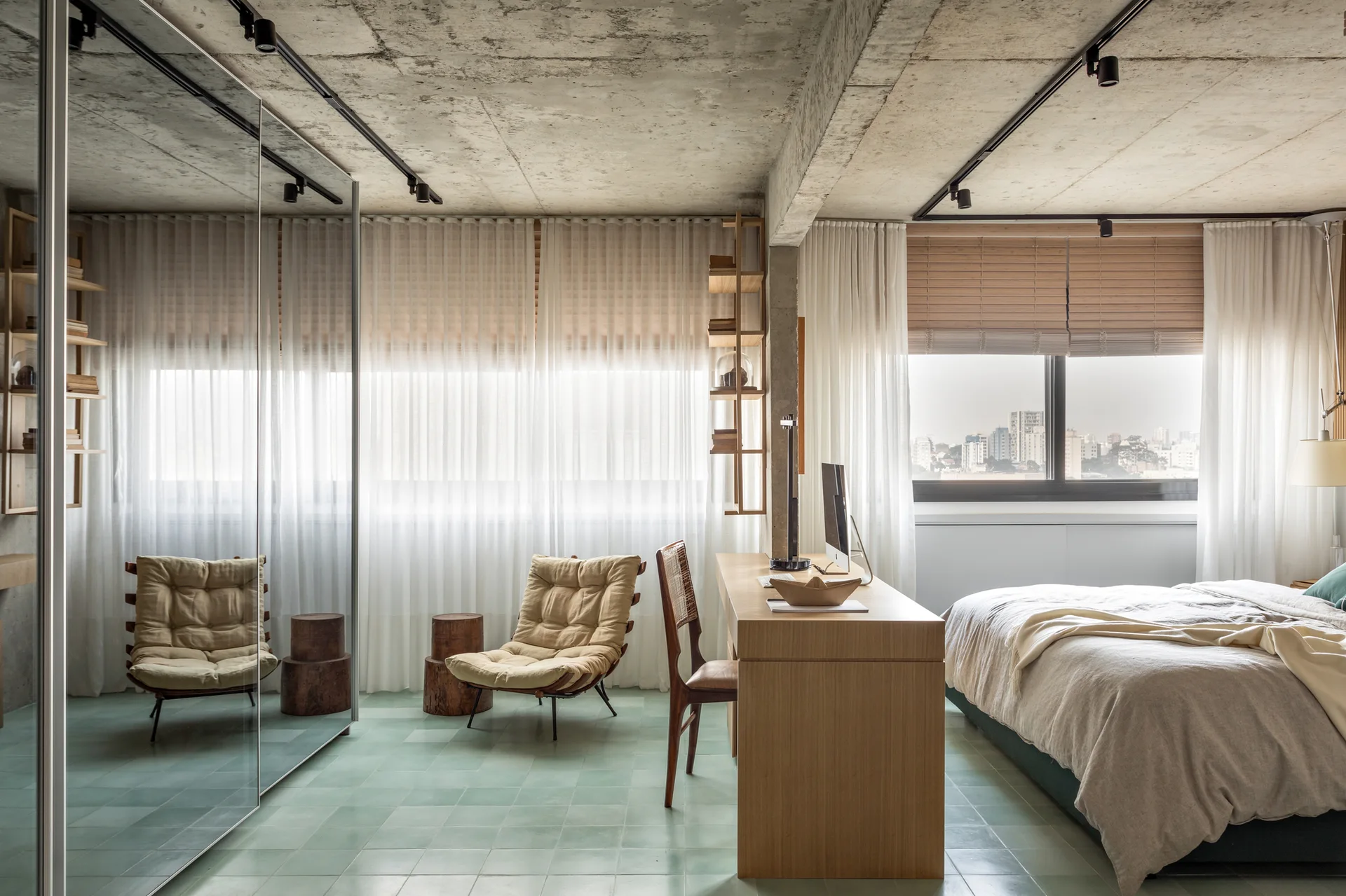 A contemporary bedroom features a green tile floor, raw concrete ceiling, wooden desk, and a large mirrored wardrobe wall.