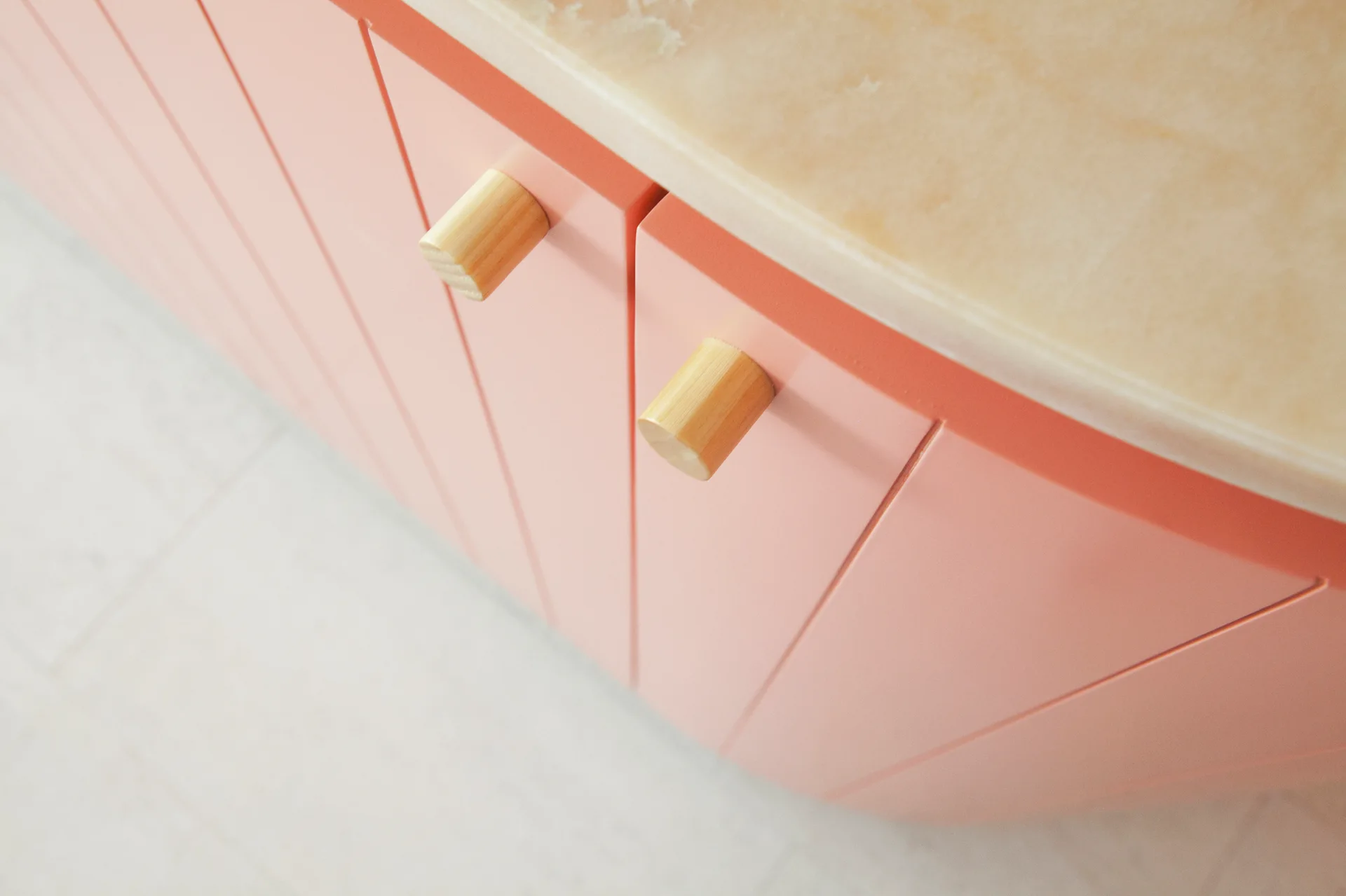 A close-up of pink cabinetry with vertical grooved panels, light wood cylindrical knobs, and a cream-colored stone countertop.