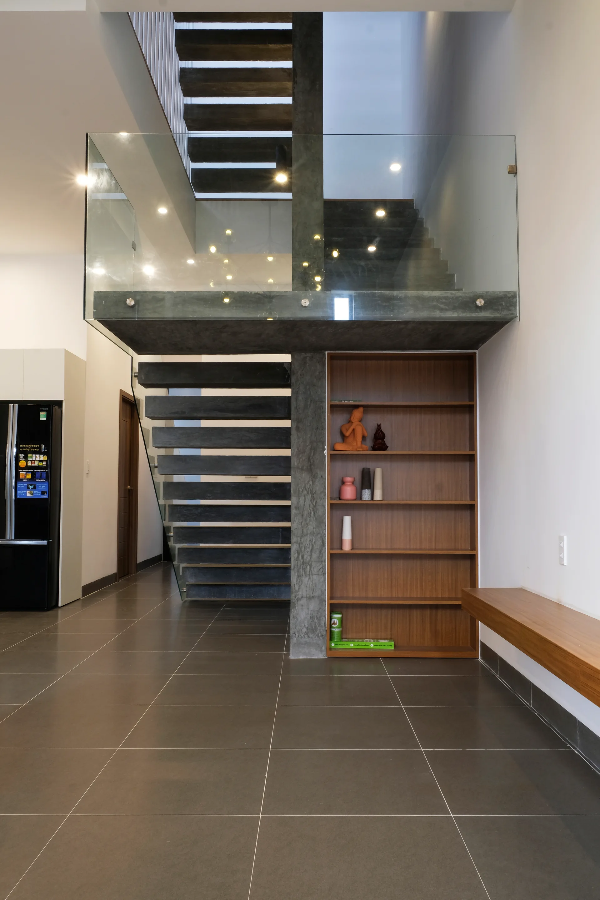 A floating concrete staircase with glass railings sits next to a built-in wooden bookshelf and dark gray floor tiles.