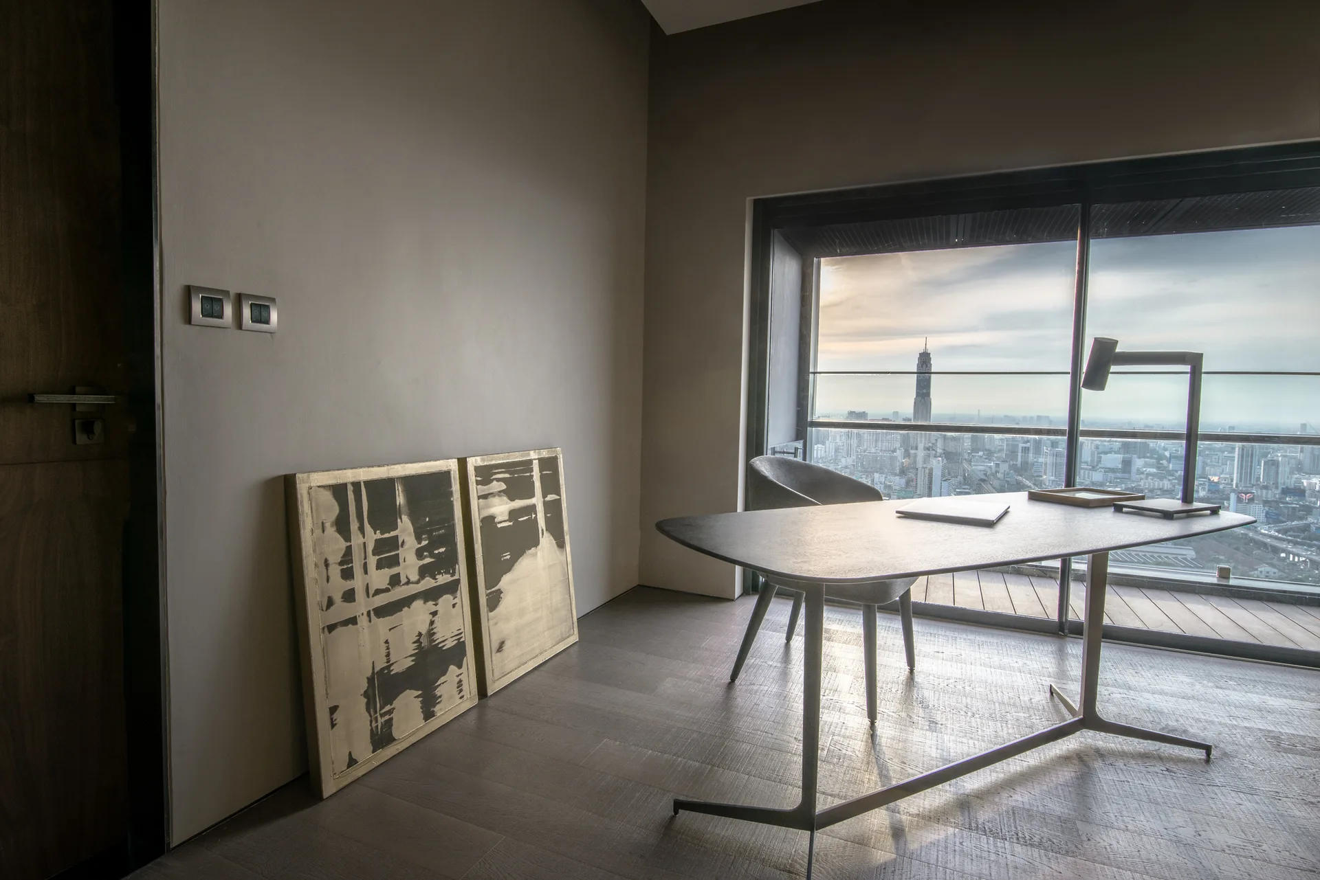 A modern office desk and chair sit in front of a large window with a cityscape view, alongside abstract art leaning against a grey wall.