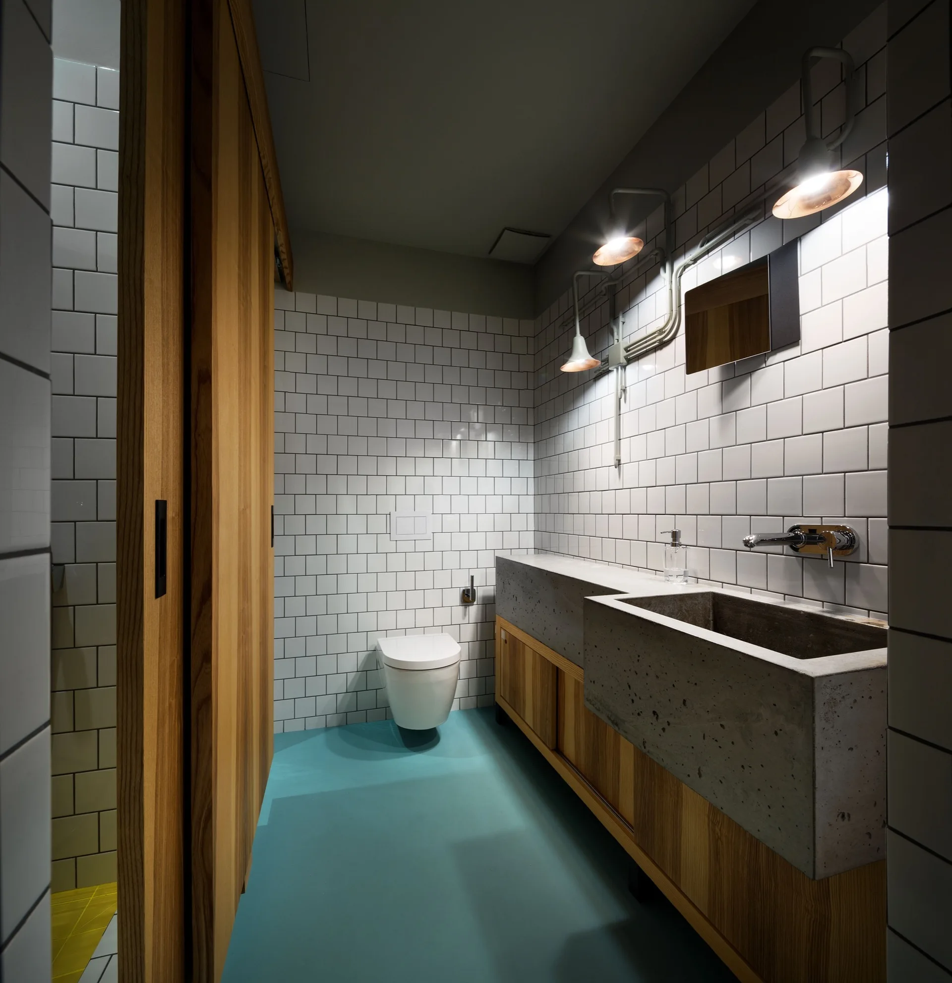 A modern bathroom featuring a concrete block vanity, a wall-mounted toilet, white grid tiles, and a vibrant turquoise floor.