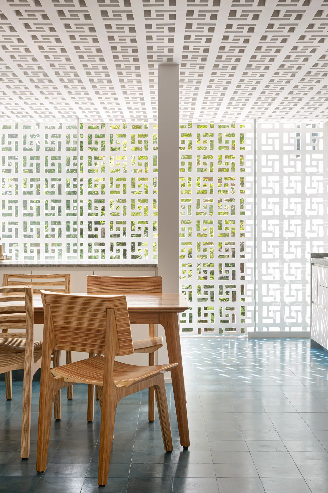 A dining room features a warm wood table and chairs set on a blue tiled floor beneath a white geometric breeze block ceiling and matching screen walls.
