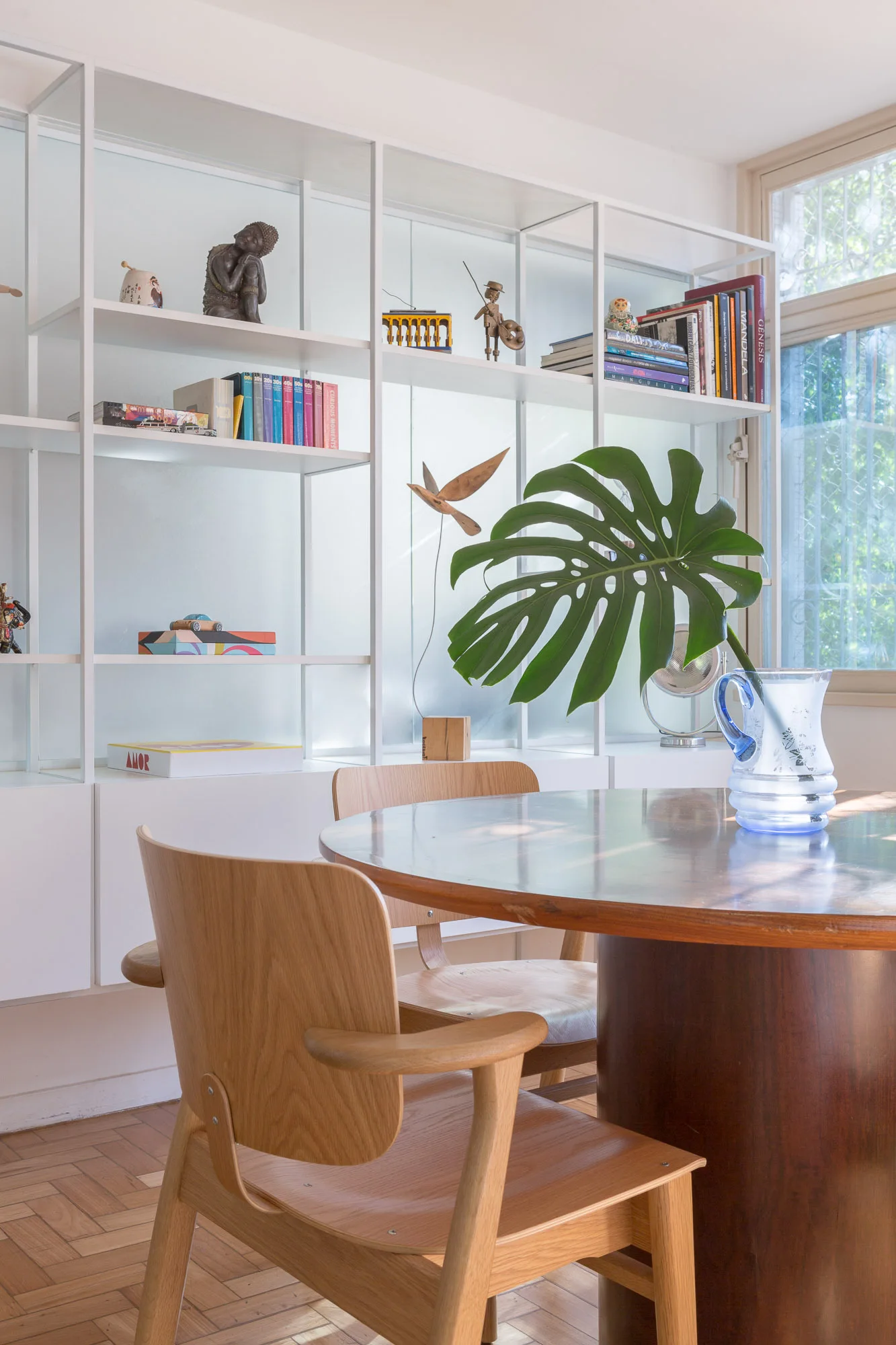 A round wood dining table and chairs are set on a parquet floor in front of a tall white grid shelving unit filled with books and decor.