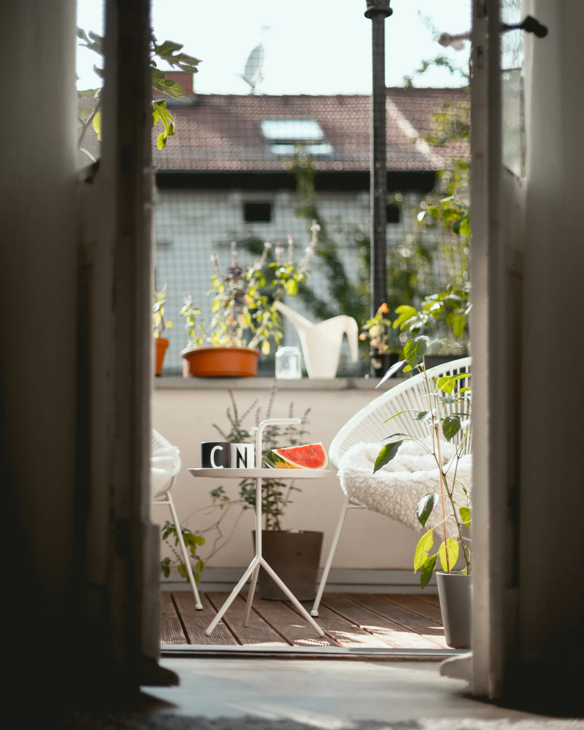 A view through an open doorway reveals a sunny wooden balcony with two white rounded chairs, a small metal table, and several potted plants.