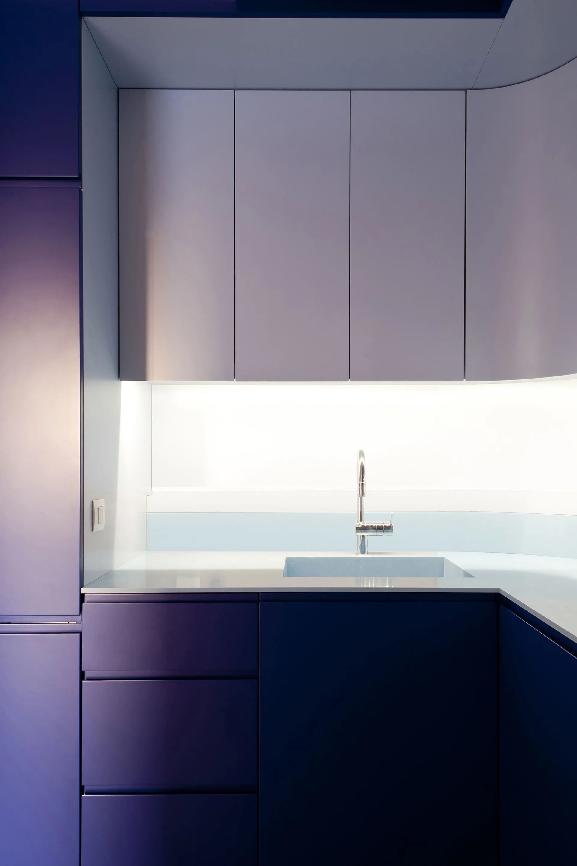 Matte purple kitchen cabinetry surrounding a seamless countertop with an integrated sink, a chrome faucet, and bright under-cabinet lighting.