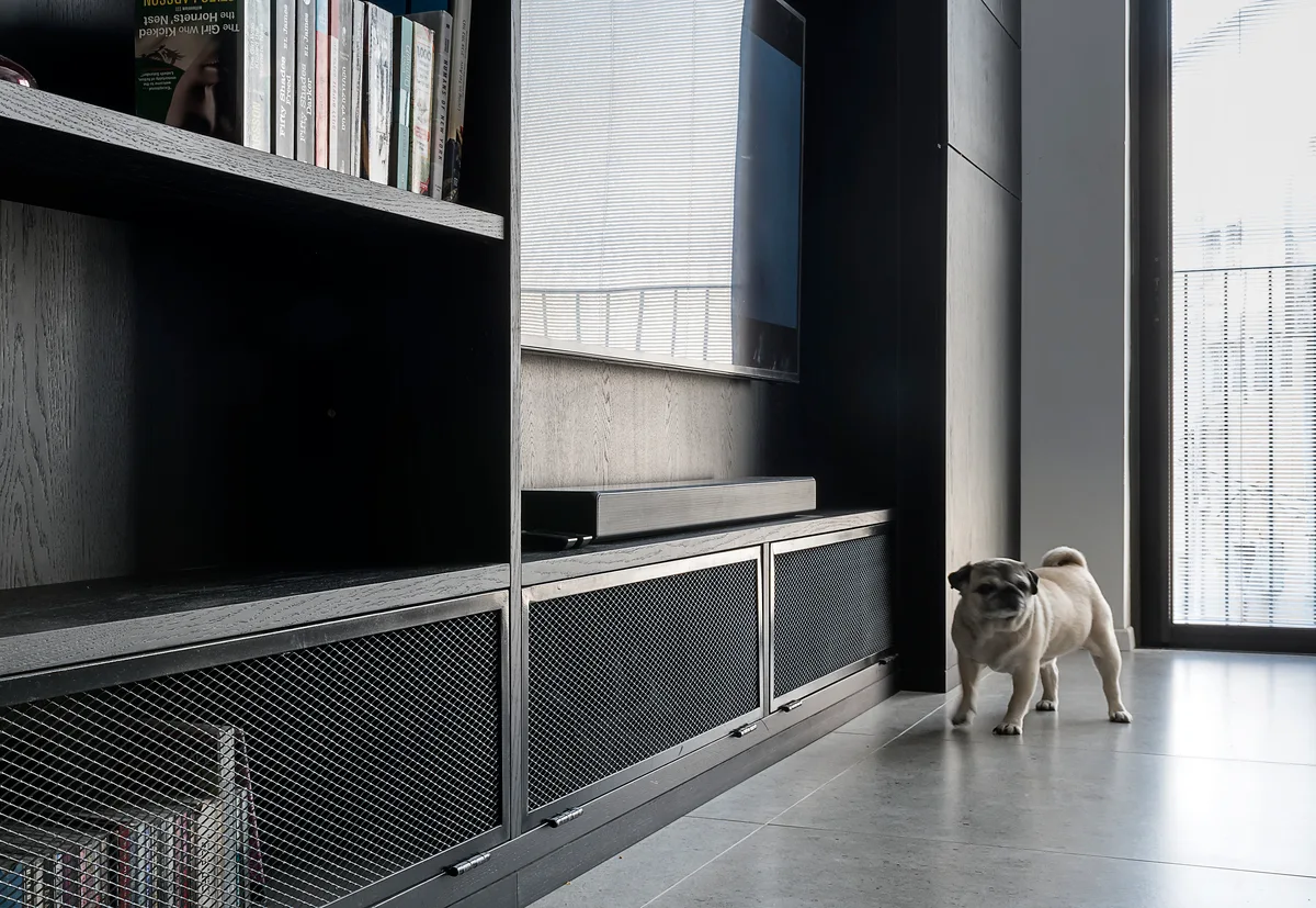 A black built-in shelving unit with wire mesh cabinet doors sits on a grey tile floor next to a window with a small dog.