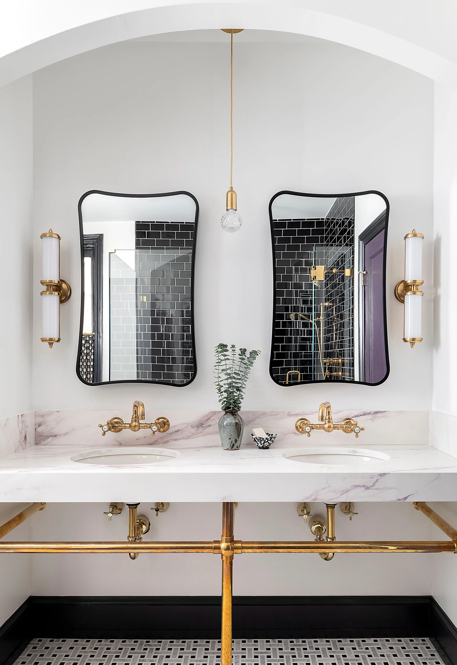 A bathroom vanity featuring a white marble countertop, two brass wall-mounted faucets, black-framed mirrors, and exposed brass legs under an arch.