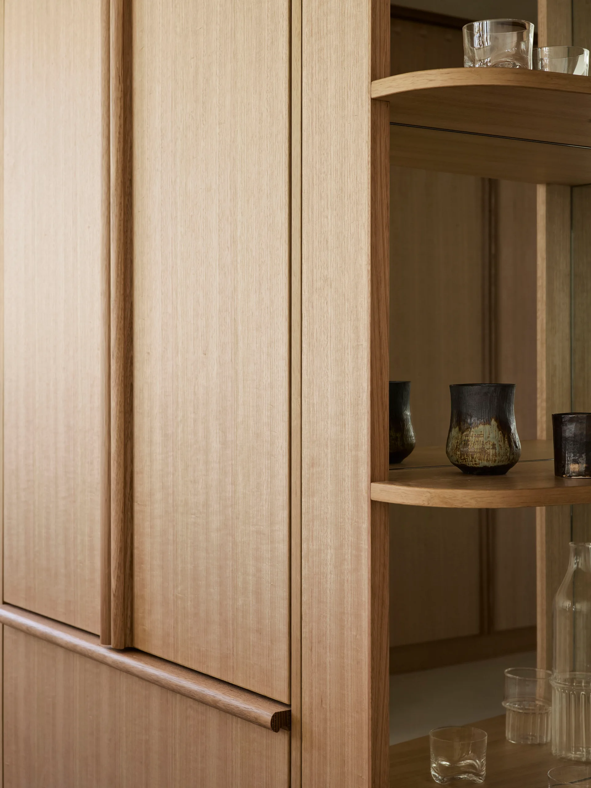 Light wood cabinet doors with vertical grain beside open curved display shelves backed with a mirror holding glassware.